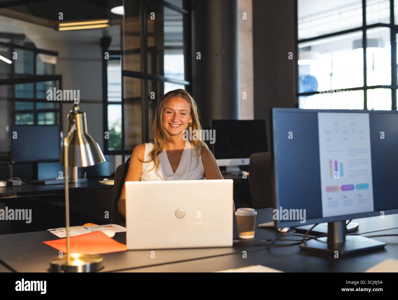 Femme professionnelle dans la trentaine examinant les graphiques sur le moniteur et l'ordinateur portable au bureau moderne Banque D'Images Femme professionnelle dans la trentaine examinant les graphiques sur le moniteur et l'ordinateur portable au bureau moderne Banque D'Images