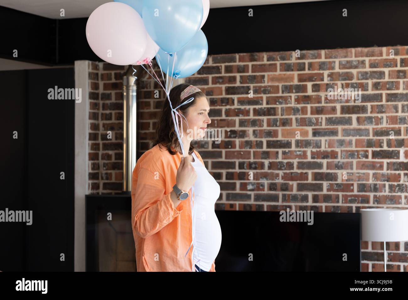 Femme enceinte tenant des ballons roses pastel et bleus dans le salon par un mur de briques, espace de copie Banque D'Images
