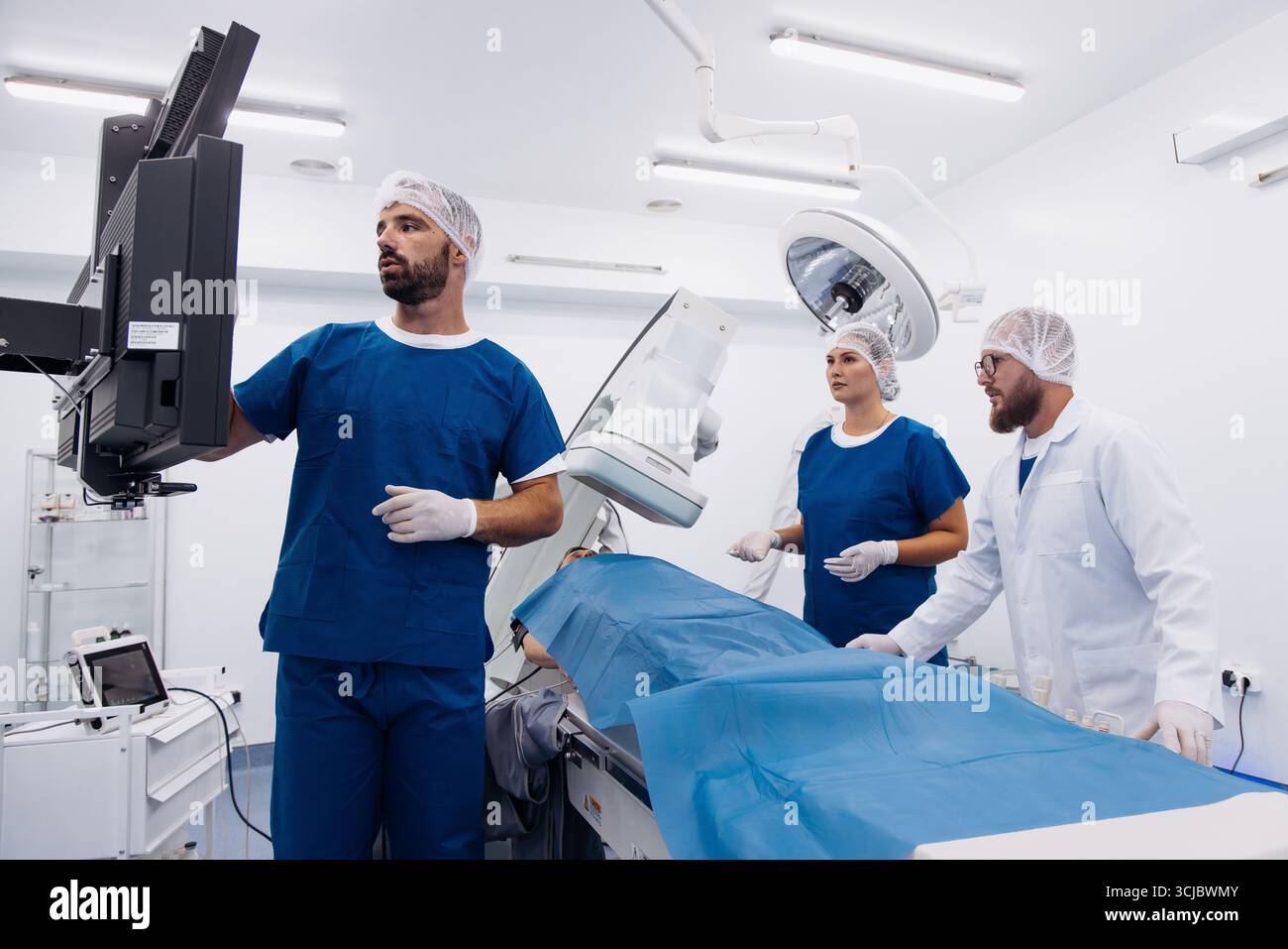L'équipe médicale en gommages bleus collabore activement dans une salle d'opération moderne, se préparant à une procédure d'angiographe avec des équipements d'imagerie avancés Banque D'Images