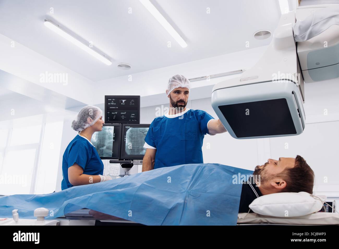Les professionnels de la santé dans les gommages bleus se préparent pour une procédure d'angiographe, avec un patient allongé sur la table d'examen et d'imagerie avancée equ Banque D'Images