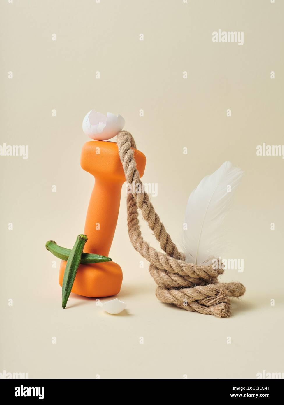 Une composition artistique de nature morte mettant en vedette un haltère orange orné d'une coquille d'œuf, entrelacé avec une corde, un gombo et une plume sur un b neutre Banque D'Images