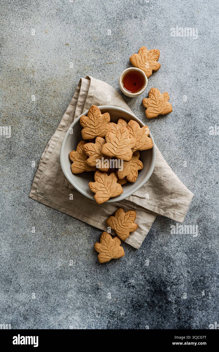 Vue de dessus de biscuits en forme de feuille d'érable dans un bol blanc sur une serviette en lin beige. Un petit bol de sirop d'érable est à proximité, posé contre un dos gris texturé Banque D'Images