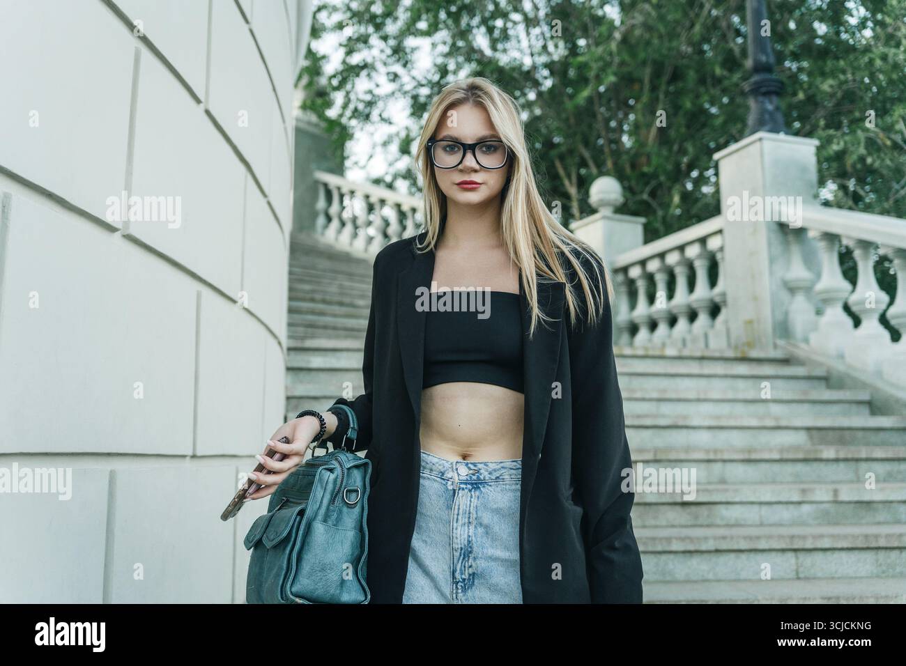 Jeune femme blonde portant une veste noire et des lunettes, debout en toute confiance à l'extérieur. Elle tient un sac bleu et un téléphone, avec des escaliers élégants et de la verdure Banque D'Images