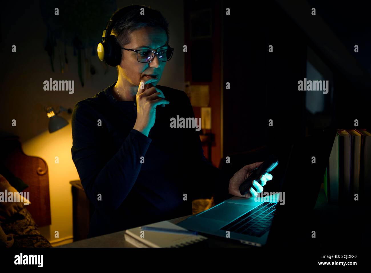 Une femme s'assoit dans son bureau à domicile la nuit avec des écouteurs, se concentrant sur l'écran de son ordinateur portable. La scène reflète le travail à distance, les outils de communication modernes Banque D'Images