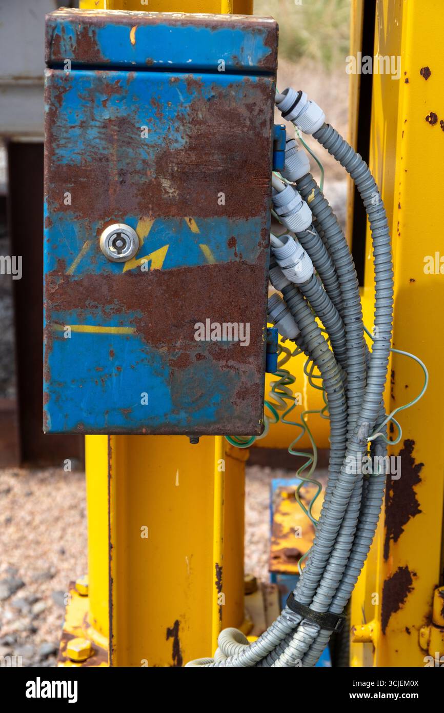 Boîte électrique en métal bleu rouillé avec câbles de conduit enroulés attachés, monté sur une structure industrielle jaune à l'extérieur, symbolisant les machines vieillissantes Banque D'Images
