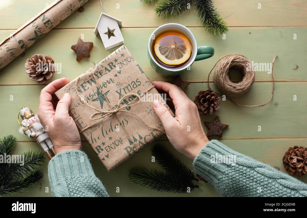 Les mains de la personne dans un pull tricoté présentent un cadeau emballé sur une table en bois rustique, entouré de décorations de vacances et une tasse de vapeur de te Banque D'Images