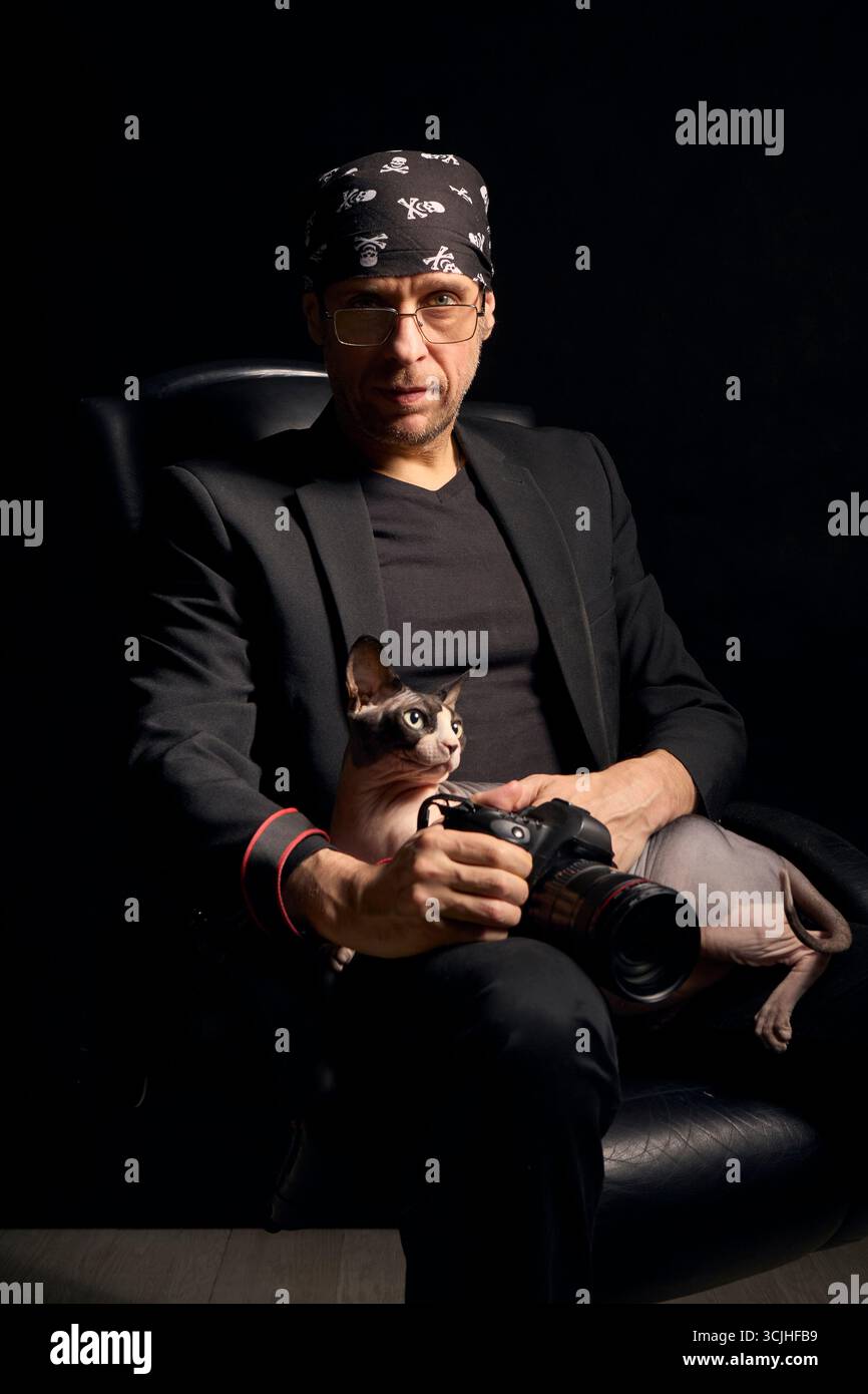 Un photographe masculin pose avec un chat Sphynx canadien dans un studio sombre. Le portrait sombre met l'accent sur la créativité, le style personnel et la compagnie Banque D'Images