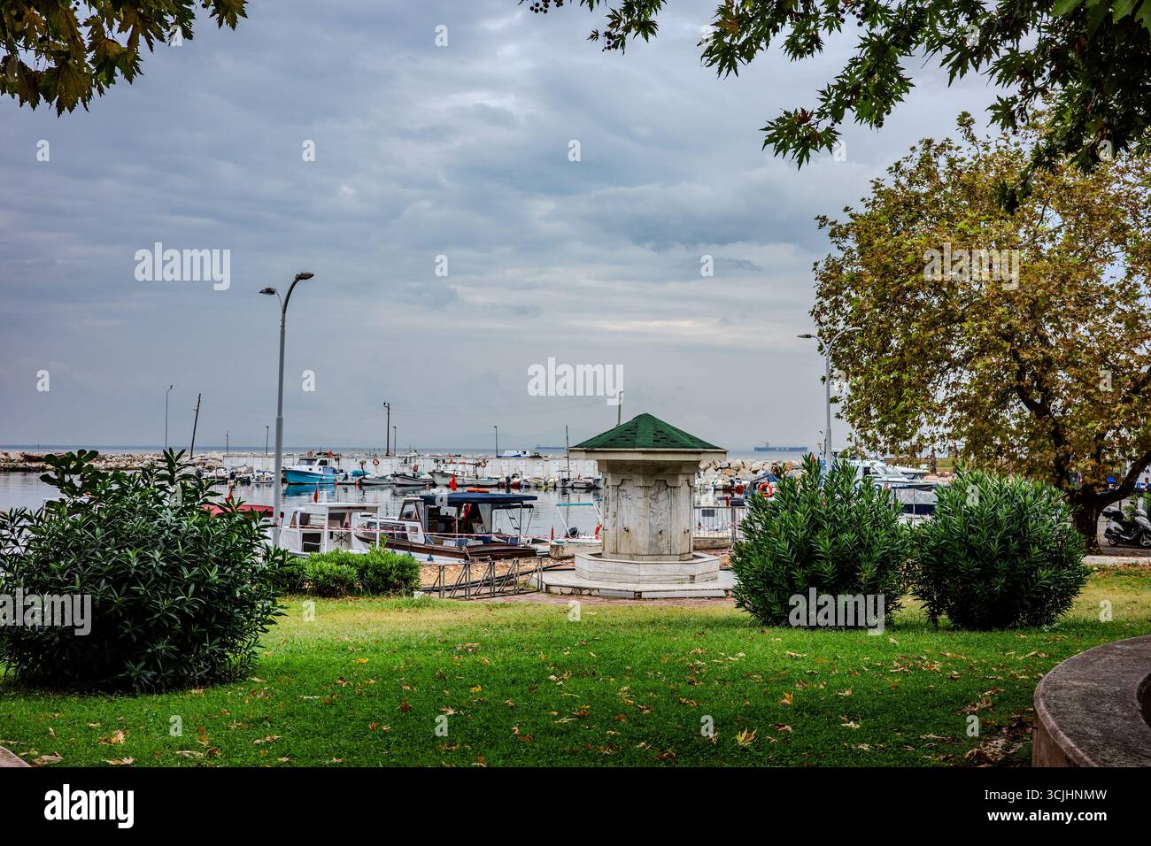 Un parc avec de la verdure et une fontaine de pierre surplombe les bateaux de pêche amarrés au port de Yalova, en Turquie, sous un ciel nuageux. Banque D'Images