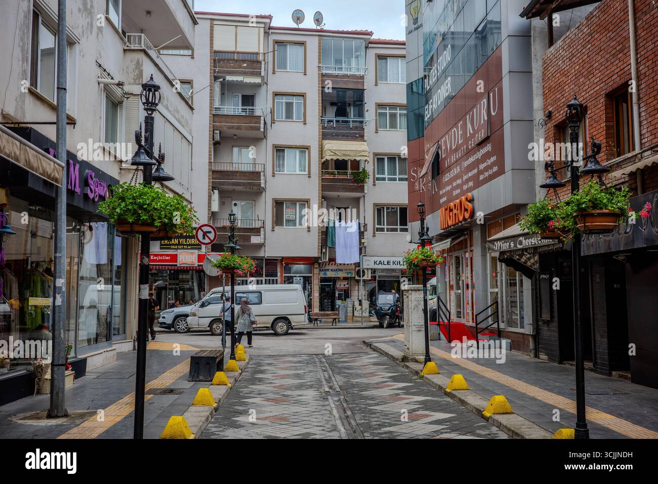 Une rue étroite bordée d'appartements résidentiels, de magasins locaux et d'un magasin Migros se trouve à Yalova, en Turquie. Paniers à fleurs suspendus et piétons a Banque D'Images