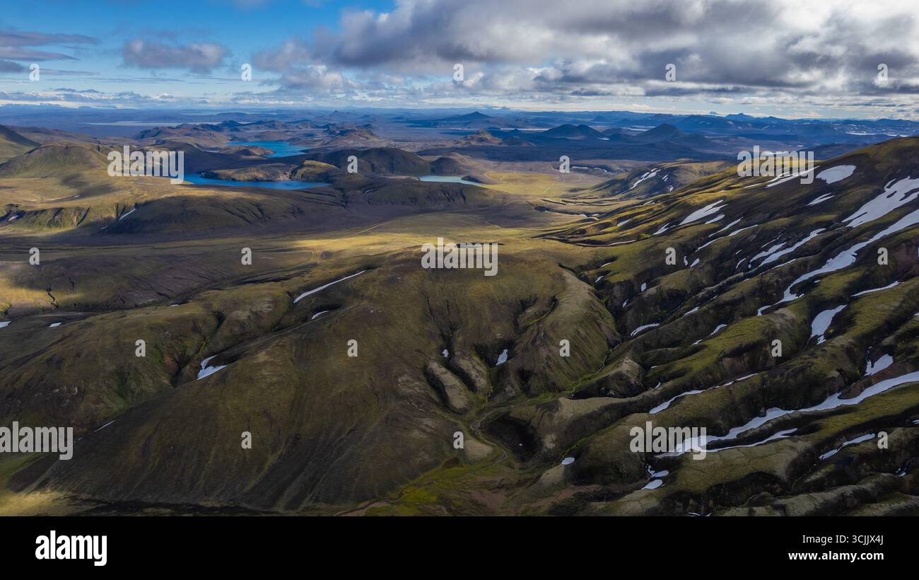 Vue aérienne des hautes terres islandaises avec des collines volcaniques ondulantes, des plages de neige et des lacs bleus sous des nuages spectaculaires dans une nature sauvage reculée Banque D'Images