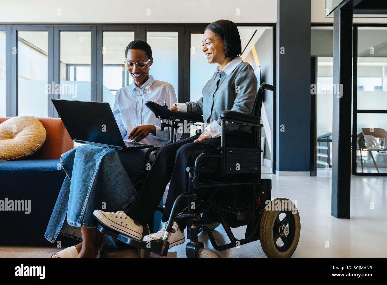 Deux femmes, dont une utilisatrice de fauteuil roulant, collaborent dans un environnement de bureau moderne, à l'aide d'un ordinateur portable. Banque D'Images