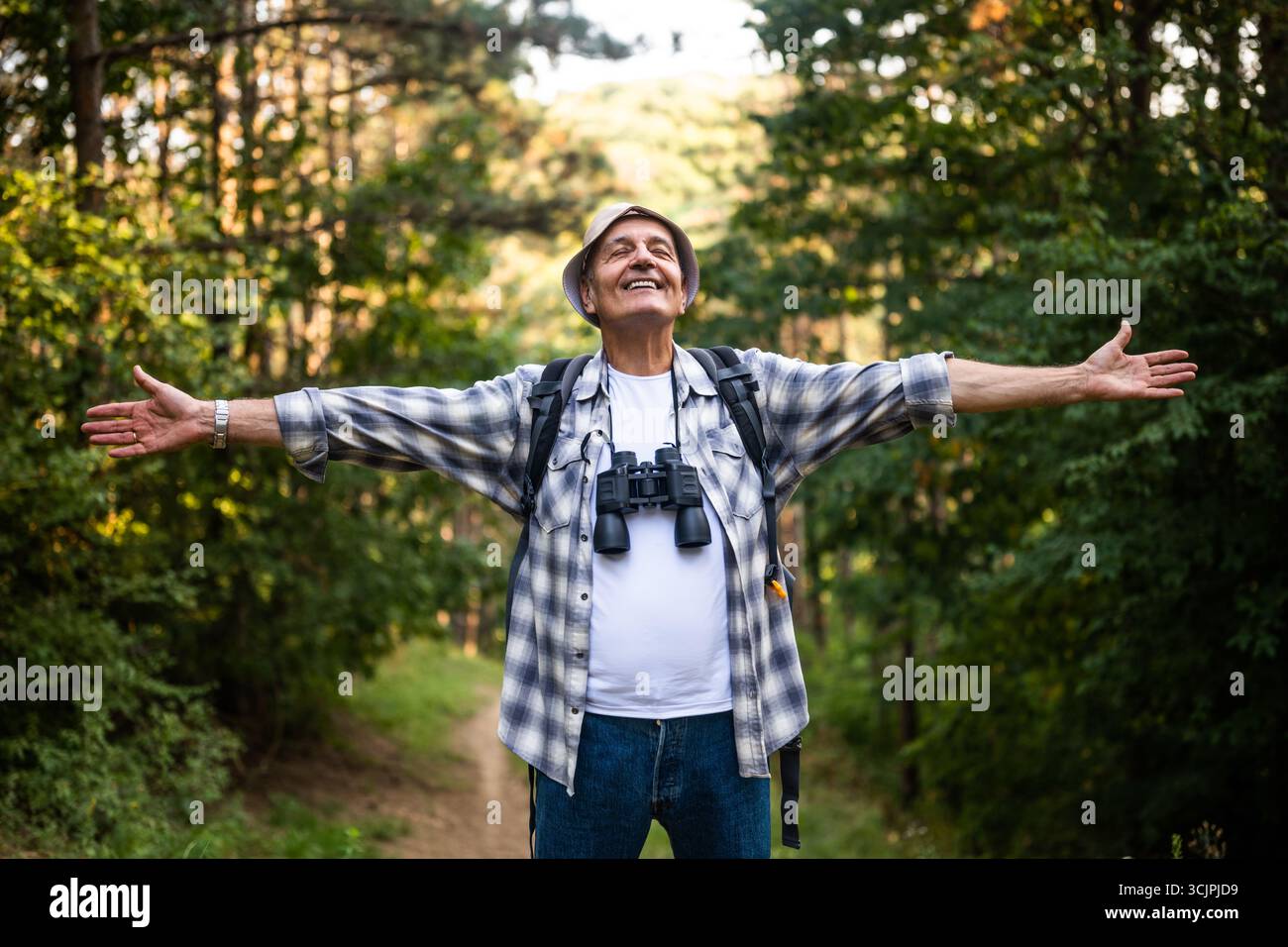 Randonneur senior heureux et insouciant avec des jumelles et un sac à dos levant les bras pendant la randonnée dans la nature. Banque D'Images