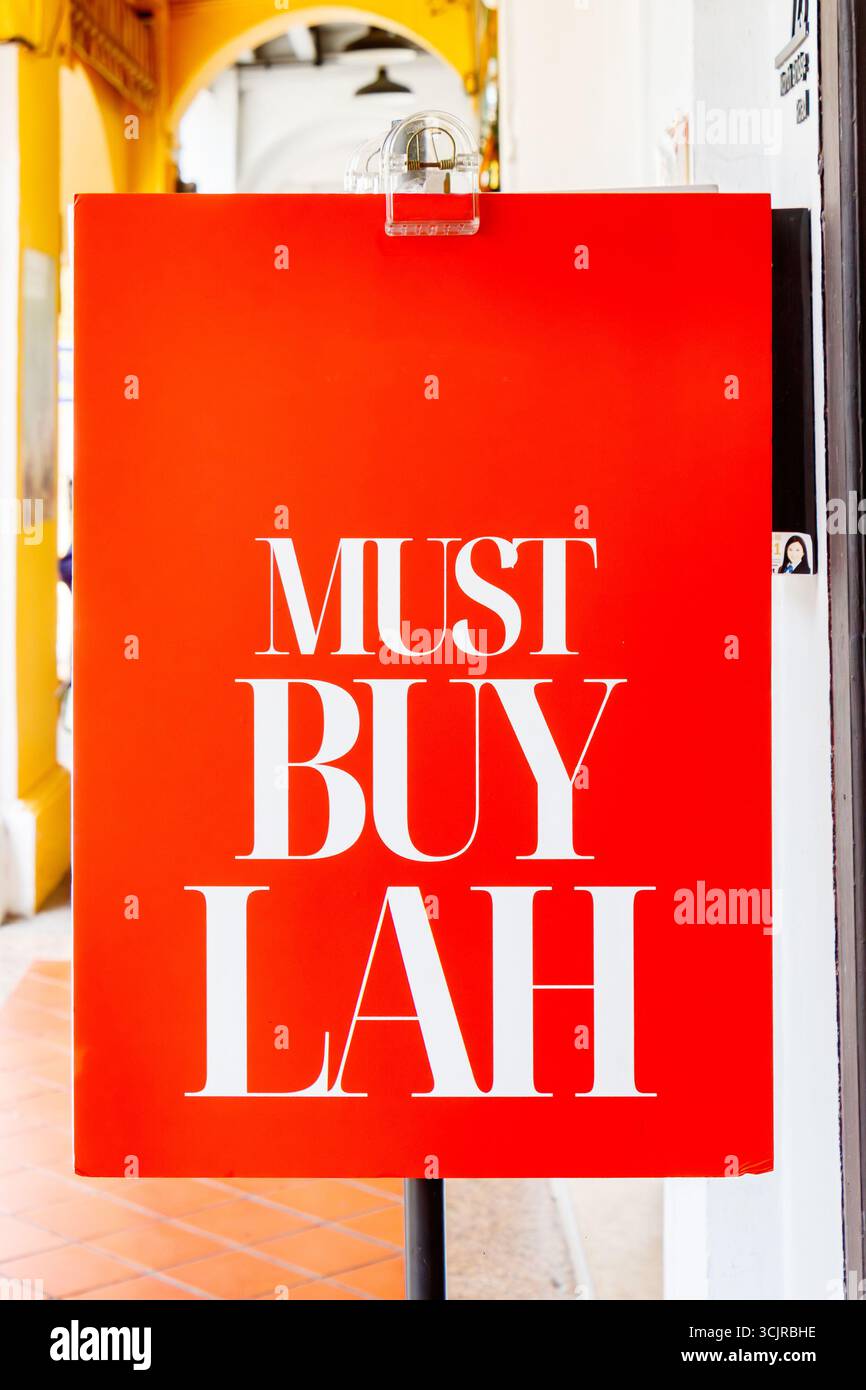 Panneau rouge en gras avec texte blanc « MUST BUY LAH » affiché dans un cadre urbain de singapour avec un fond éclatant. Banque D'Images