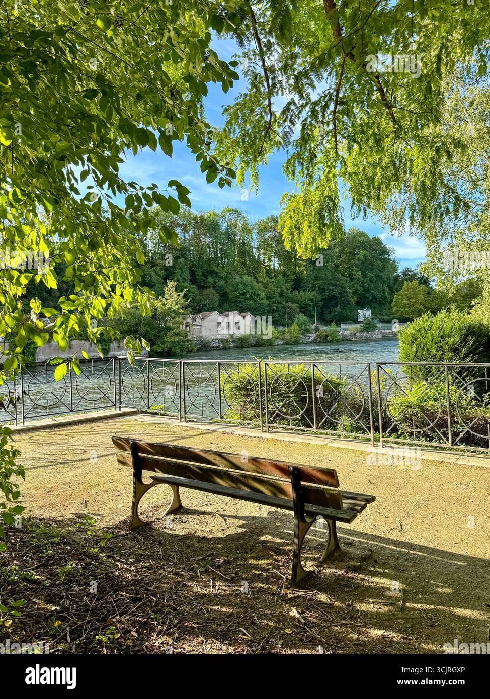 Banc en bois surplombant le calme fleuve Doubs. en arrière-plan, l'ancienne usine historique des forges à audicourt, entourée d'arbres verdoyants - Image de stock capturée avec un smartphone