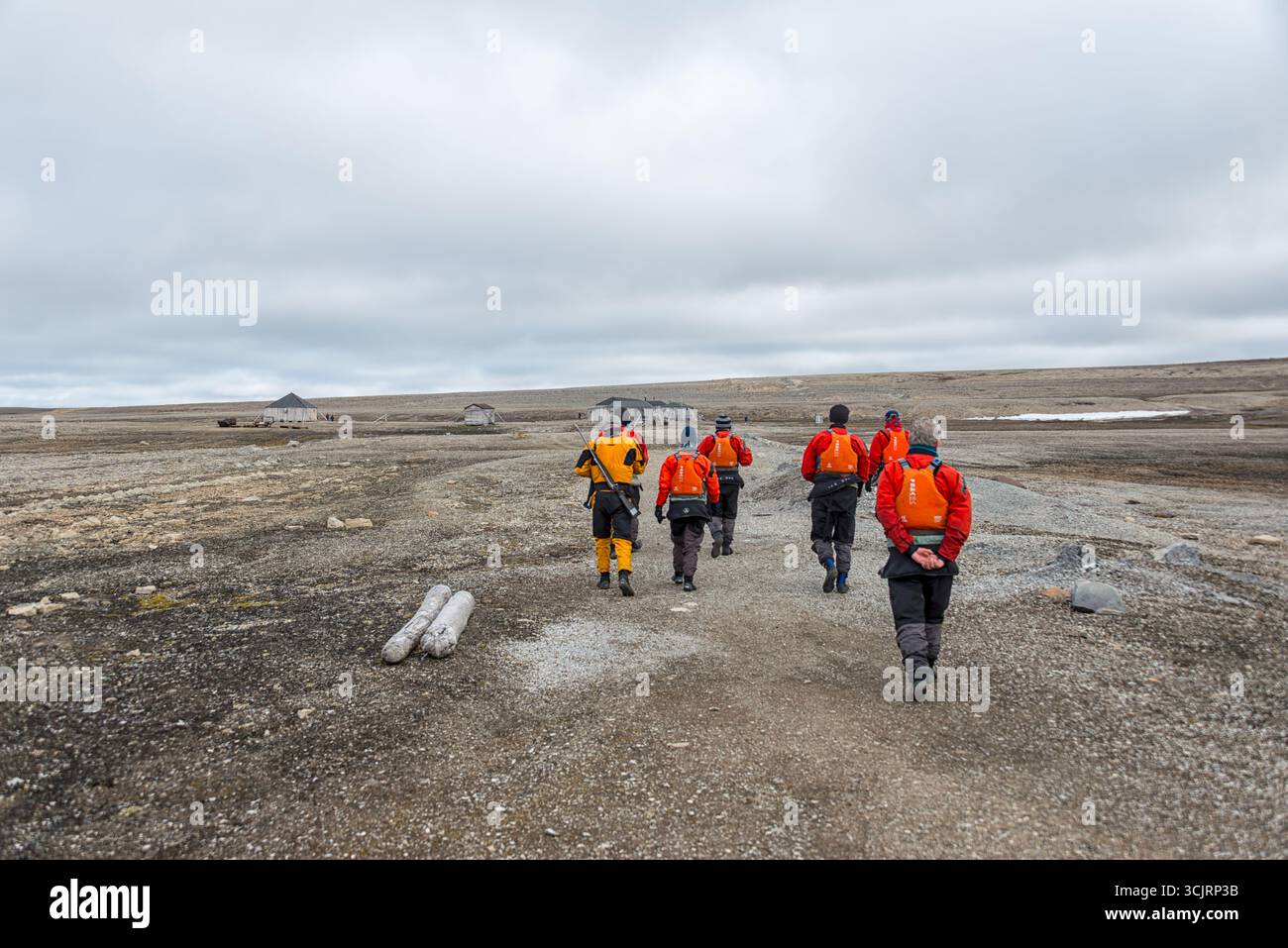 Touristes en kayak polaire marchent vers Kinnvika Station Nordaustlandet Svalbard // NORDEAUSTLANDET, Svalbard — les touristes en gilets de kayak orange vif et en équipement imperméable marchent le long d'un chemin de gravier vers un groupe de bâtiments en bois sur la toundra arctique stérile et rocheuse. Au loin, plusieurs structures, dont un bâtiment plus grand au toit sombre, sont visibles sous un ciel couvert. Cette scène capture les visiteurs arrivant à la station Kinnvika, un centre de recherche éloigné créé en 1957 dans le cadre de l’année géophysique internationale. La station représente d'importantes SCI internationales Banque D'Images