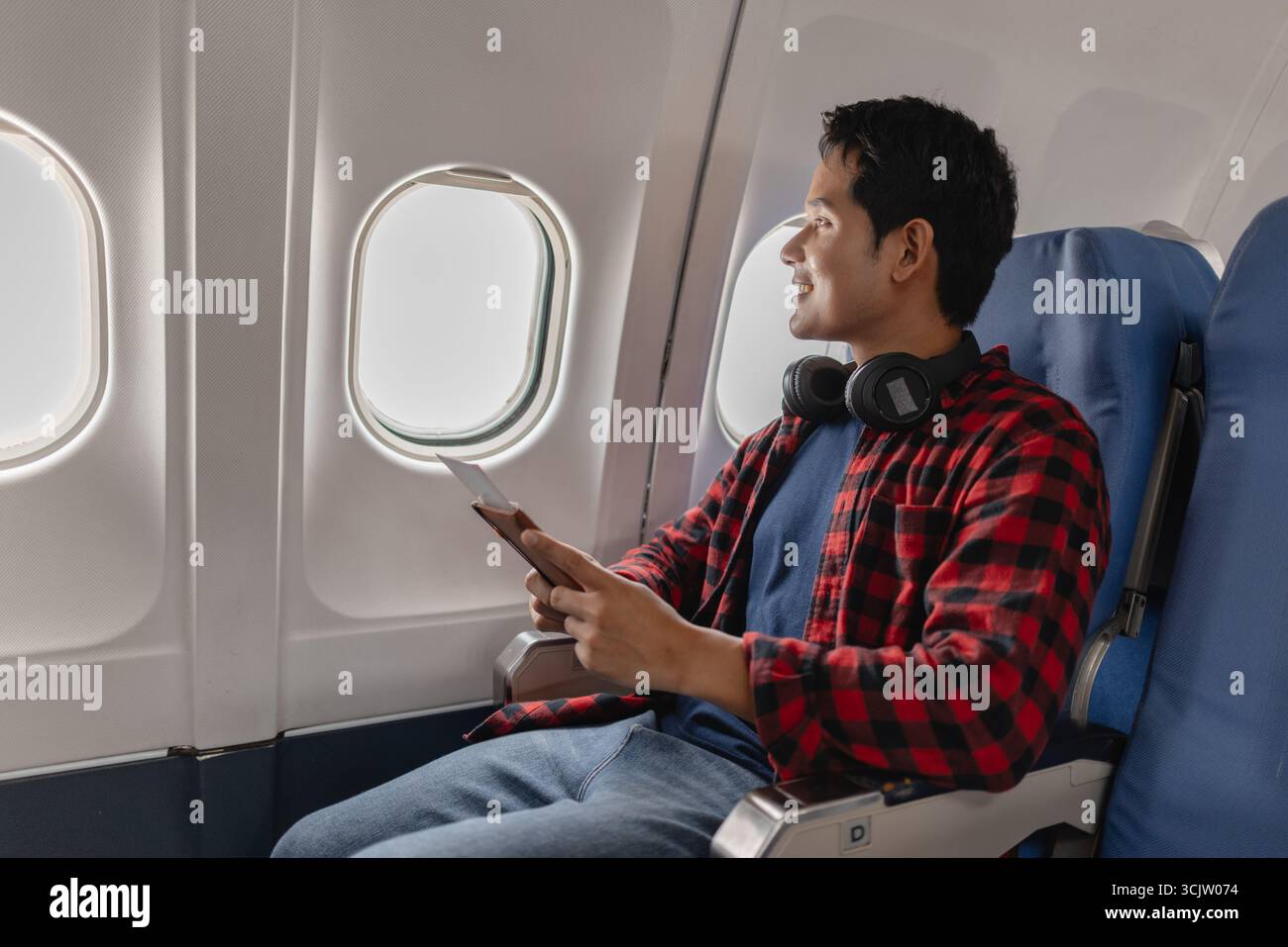 Un jeune homme est assis sur un siège côté fenêtre à l'intérieur d'un avion, tenant son passeport et sa carte d'embarquement tout en regardant par la fenêtre avec un sourire plein d'espoir. Il voit Banque D'Images