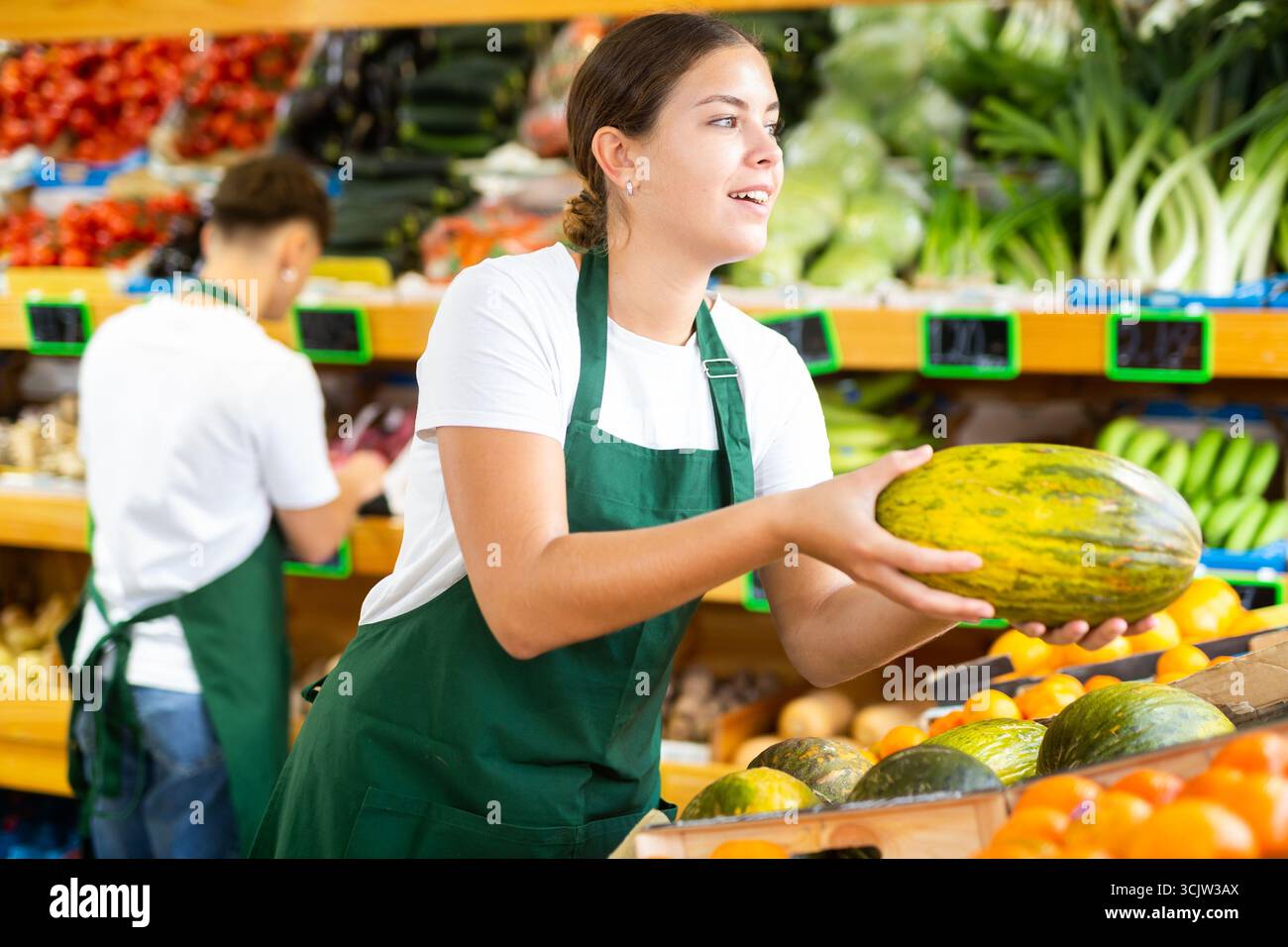 Jeune femme joyeuse ouvrière de supermarché dans le tablier tenant le melon mûr dans la section des fruits Banque D'Images