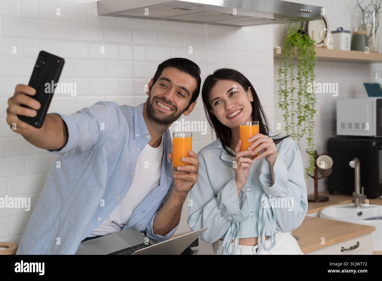 Jeune couple assis ensemble dans la cuisine moderne, souriant et prenant selfie avec smartphone tout en tenant des verres de jus d'orange, concept de bonheur, l Banque D'Images