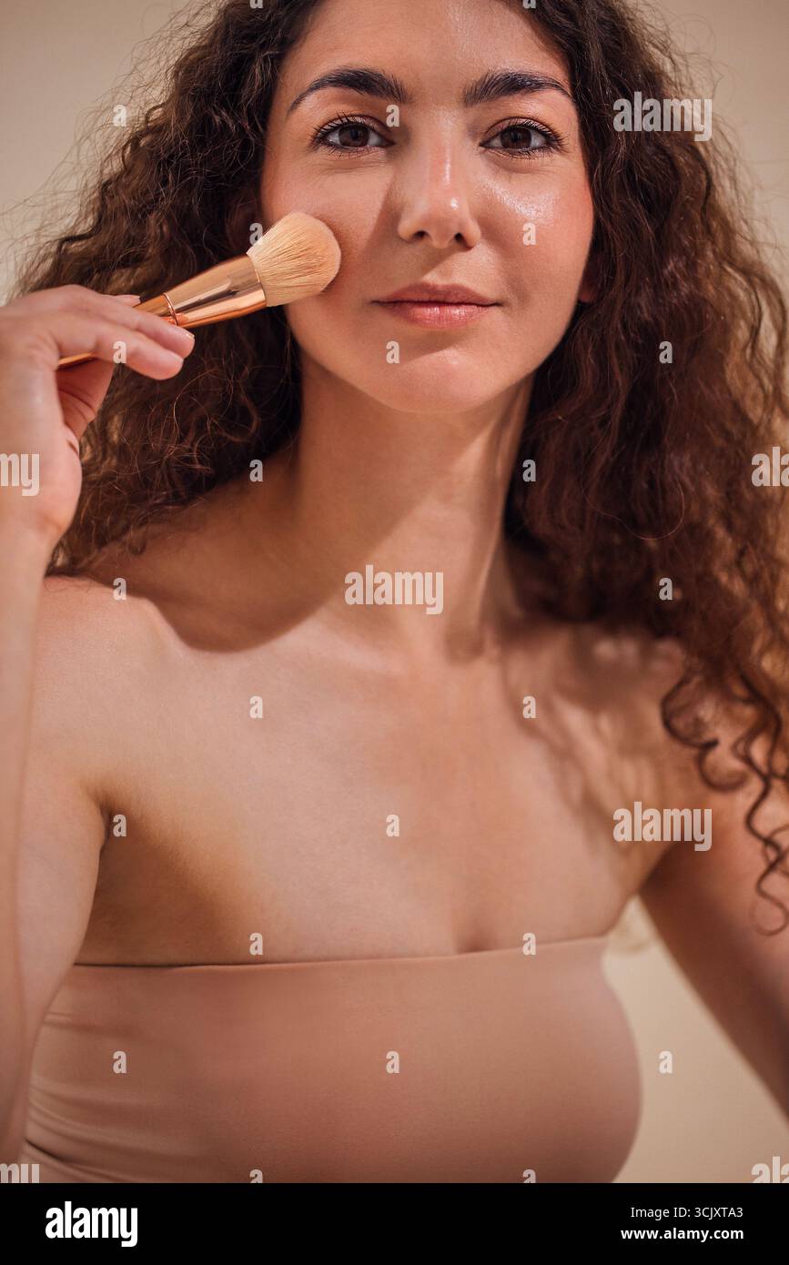 Femme appliquant le maquillage avec un pinceau sur sa joue, soulignant la routine beauté et l'auto-soin dans un environnement serein avec un éclairage doux et col neutre Banque D'Images