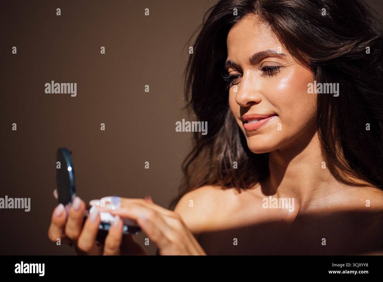 Femme aux longs cheveux foncés applique le maquillage à l'aide d'un petit miroir, soulignant sa routine beauté dans une atmosphère chaleureuse et accueillante avec un éclairage doux Banque D'Images