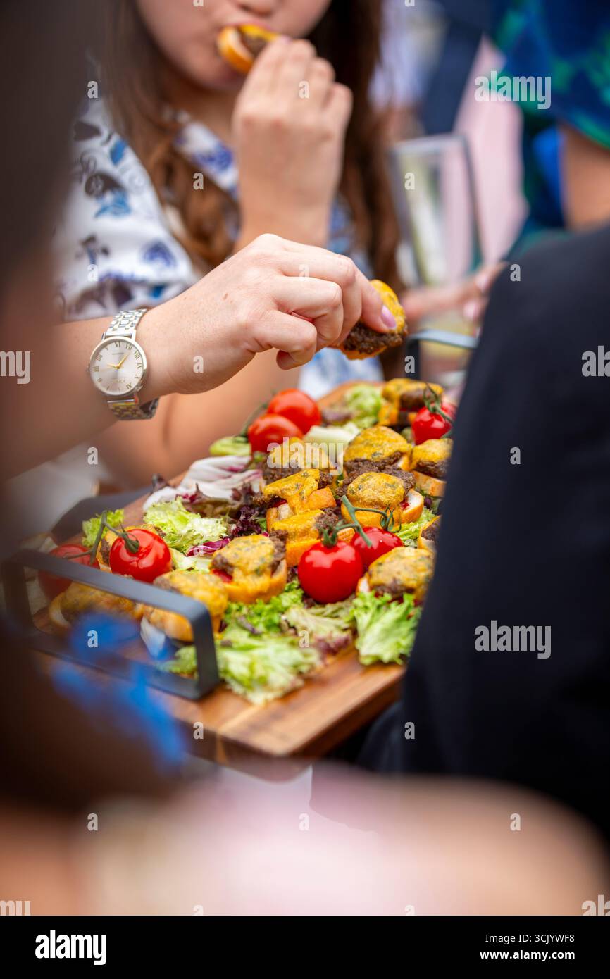 Vous pourrez déguster un plateau de cheeseburgers miniatures accompagné d'un assortiment de canapés savoureux, dont des tomates cerises, des légumes verts frais et d'autres morceaux Banque D'Images