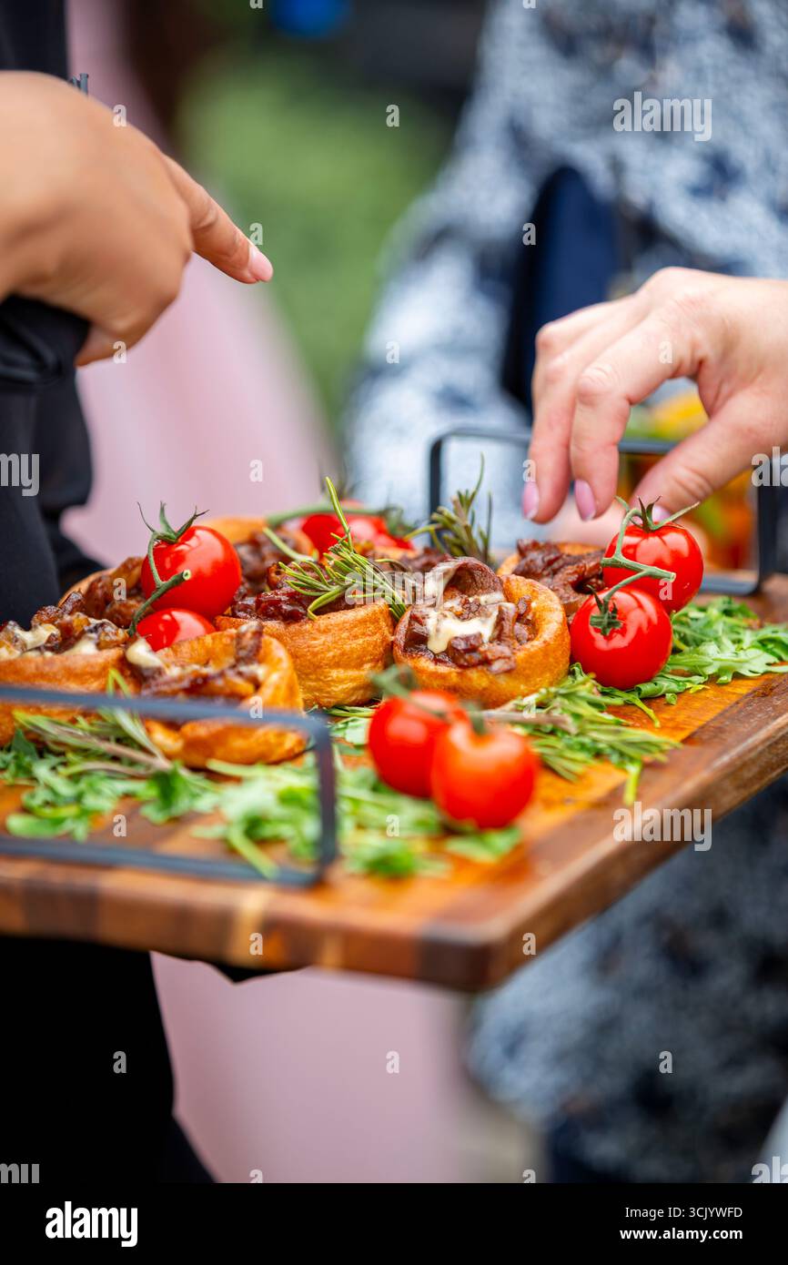 Serveur présentant une sélection de canapés élégants sur une planche de bois, avec une variété de bouchées salées garnies d'herbes fraîches et de tomates cerises Banque D'Images