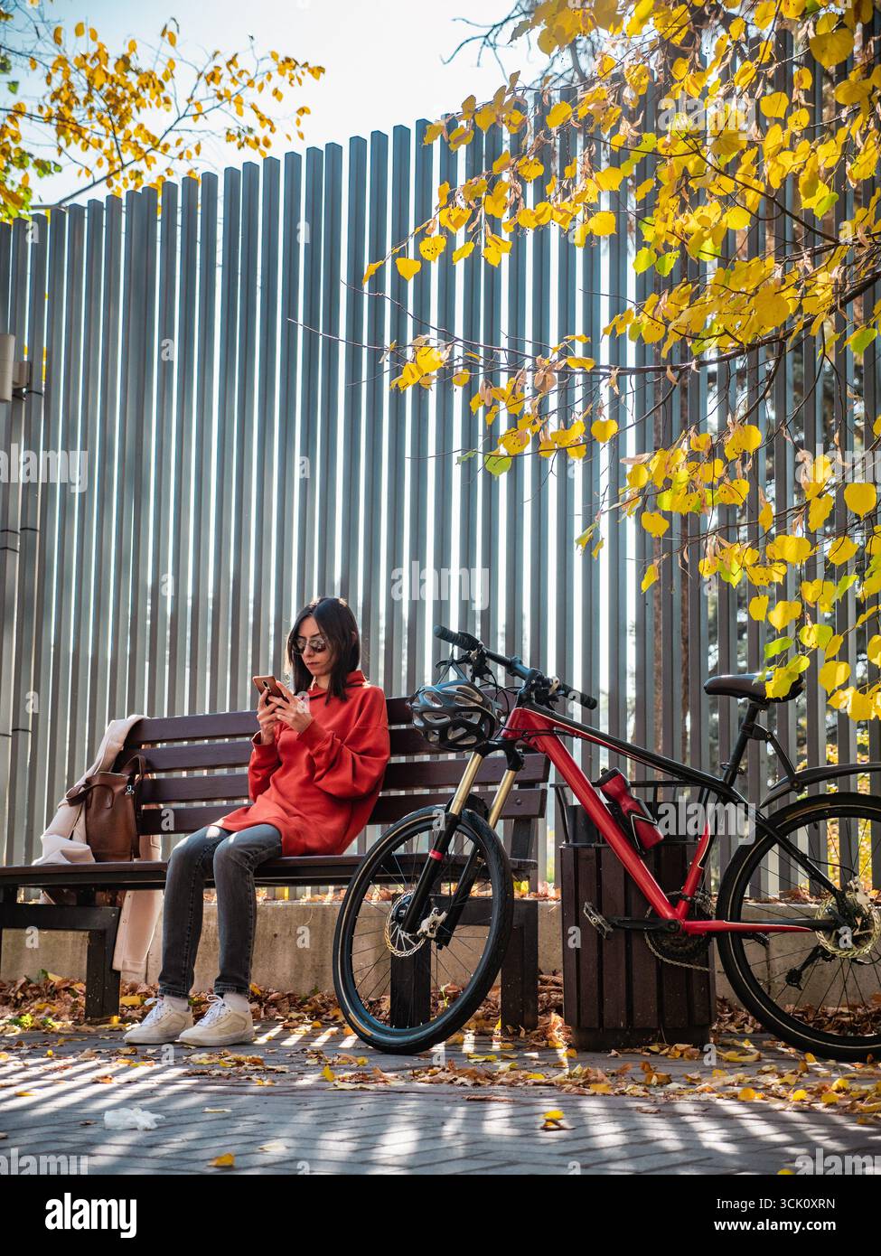 Une fille est assise sur un banc dans un parc, tenant un smartphone dans ses mains, avec son vélo debout à proximité. Temps de loisirs actif dans la ville en automne Banque D'Images