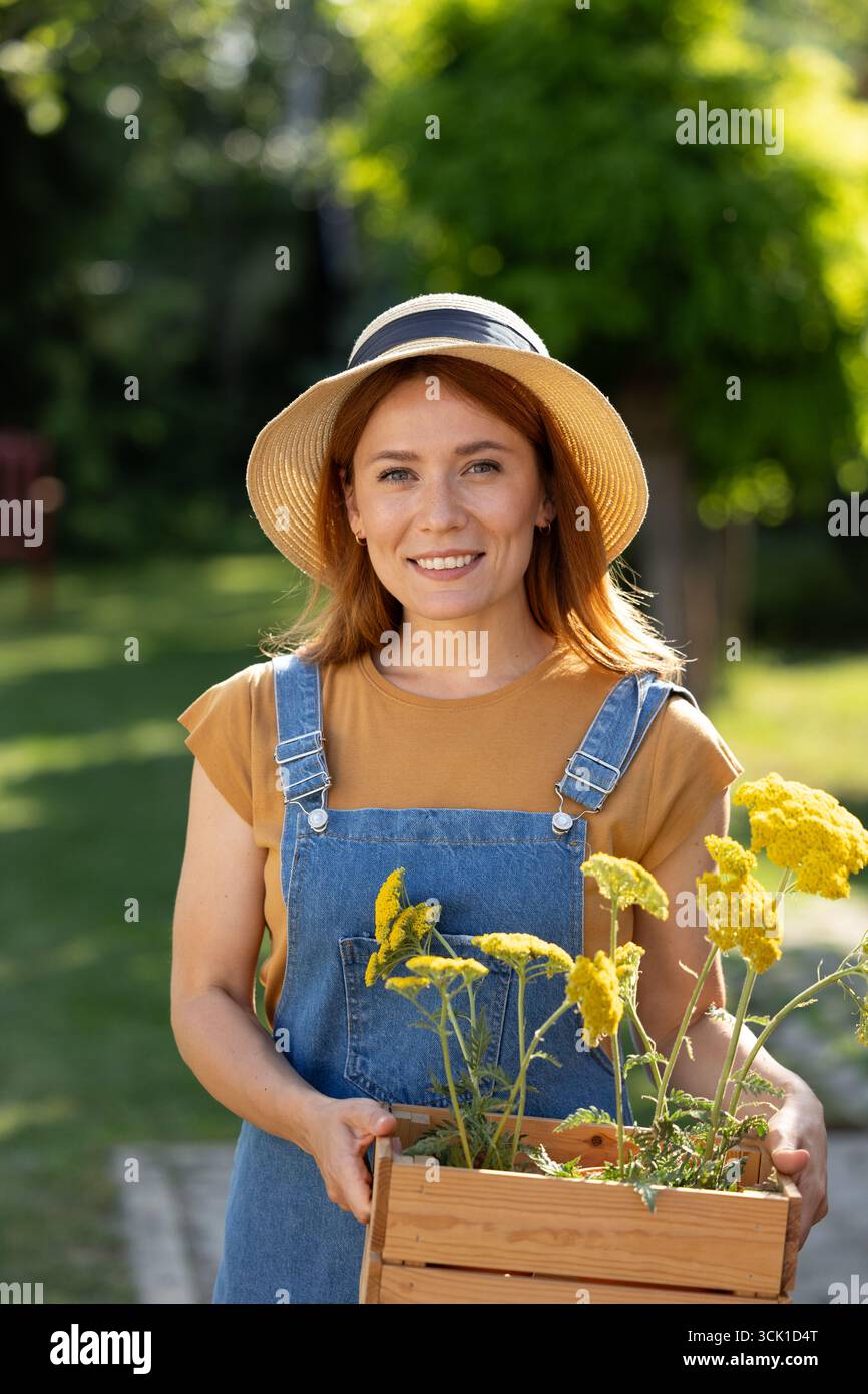 Portrait de jolie femme porte une boîte en bois de fleurs jaunes tout en profitant du jardinage dans un parc verdoyant le jour ensoleillé Banque D'Images
