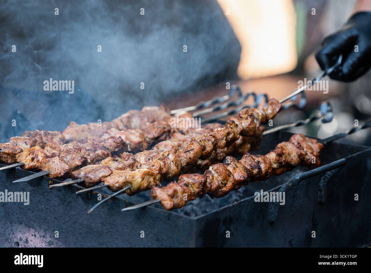 Gros plan d'une personne grillant des brochettes de viande de porc sur un barbecue, portant un gant noir Banque D'Images
