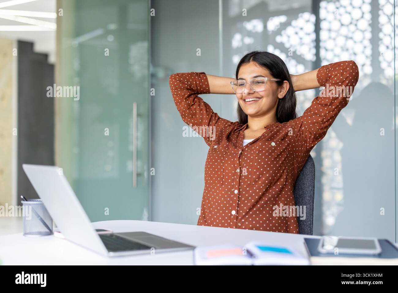 Femme d'affaires souriante se relaxant à son bureau avec les mains derrière la tête, profitant d'une pause bien méritée après avoir terminé un travail réussi sur son ordinateur portable dans un bureau lumineux et moderne Banque D'Images