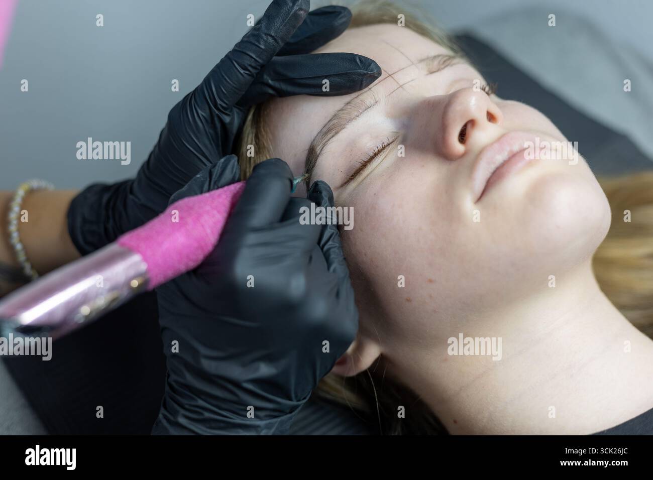Un technicien de beauté dessine soigneusement la forme des sourcils sur la peau d'un client à l'aide d'un outil de tatouage. La cliente est allongée sur une table, les yeux fermés, détendue dure Banque D'Images