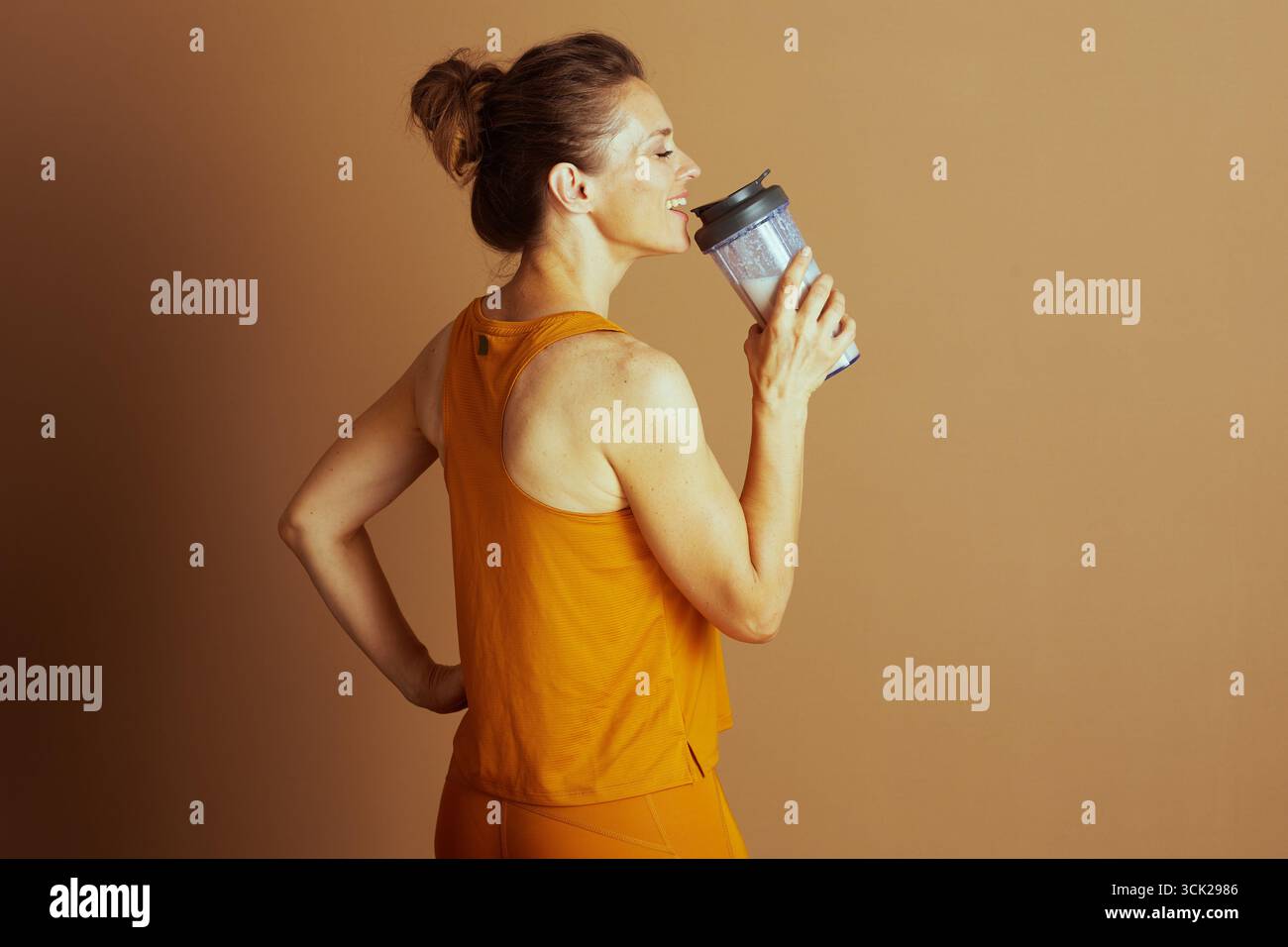 Une femme souriante dans une tenue athlétique orange se tient en profil latéral, buvant dans une bouteille de mélangeur. Sa pose avec la main sur la hanche suggère un R post-entraînement Banque D'Images
