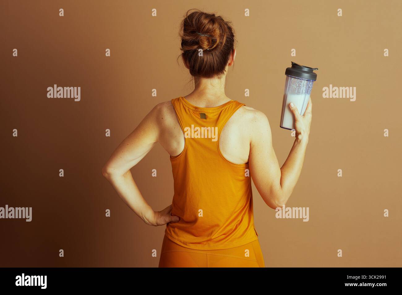 Une femme dans un débardeur doré dos nageur et leggings assortis est représentée de derrière, tenant une bouteille shaker. Sa pose avec la main sur la hanche met en évidence un focus Banque D'Images
