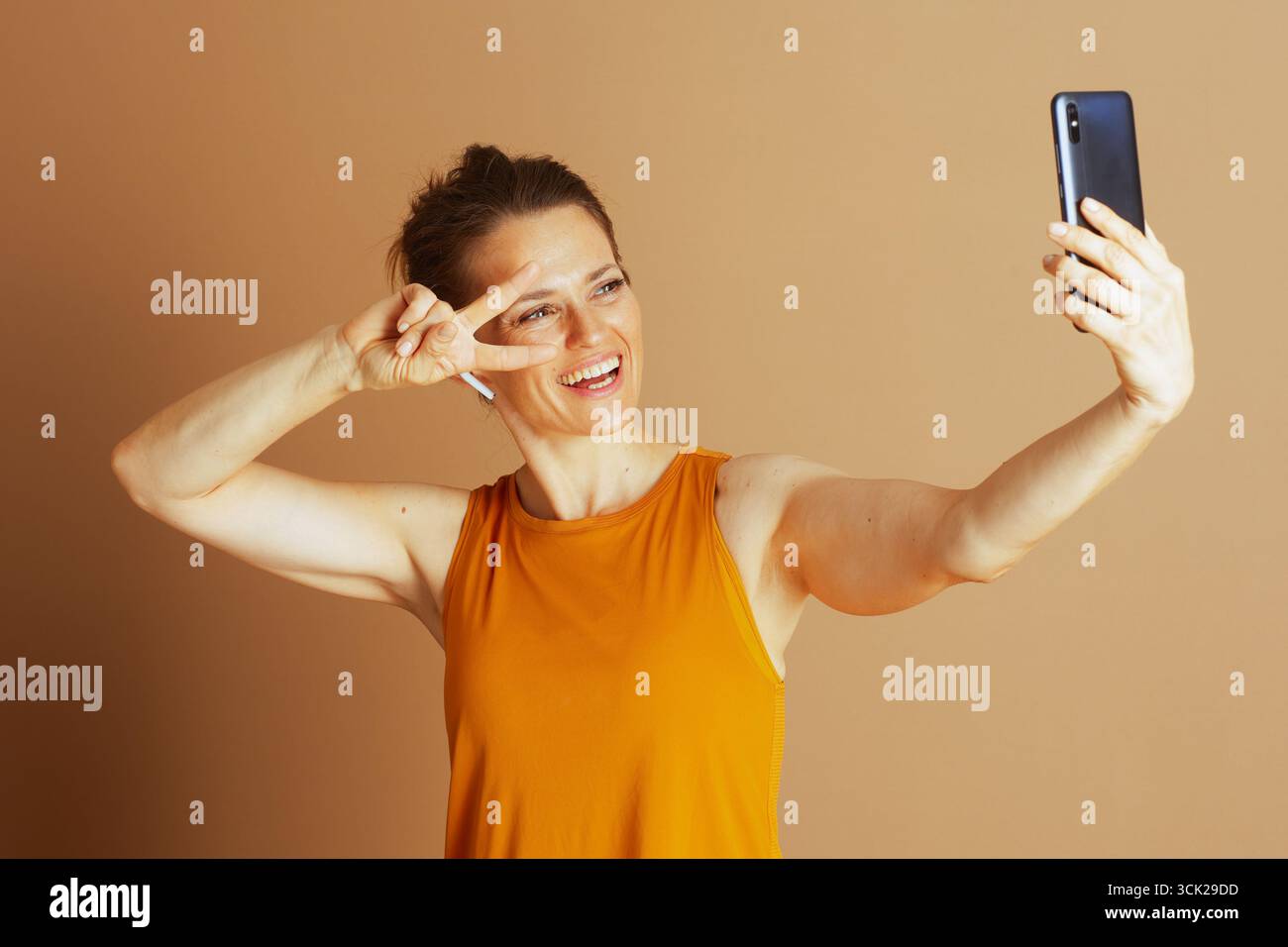 Une femme souriante dans un haut orange et un écouteur fait un signe de paix tout en prenant un selfie avec son smartphone. Elle rayonne de gaieté et décontractée, moderne Banque D'Images