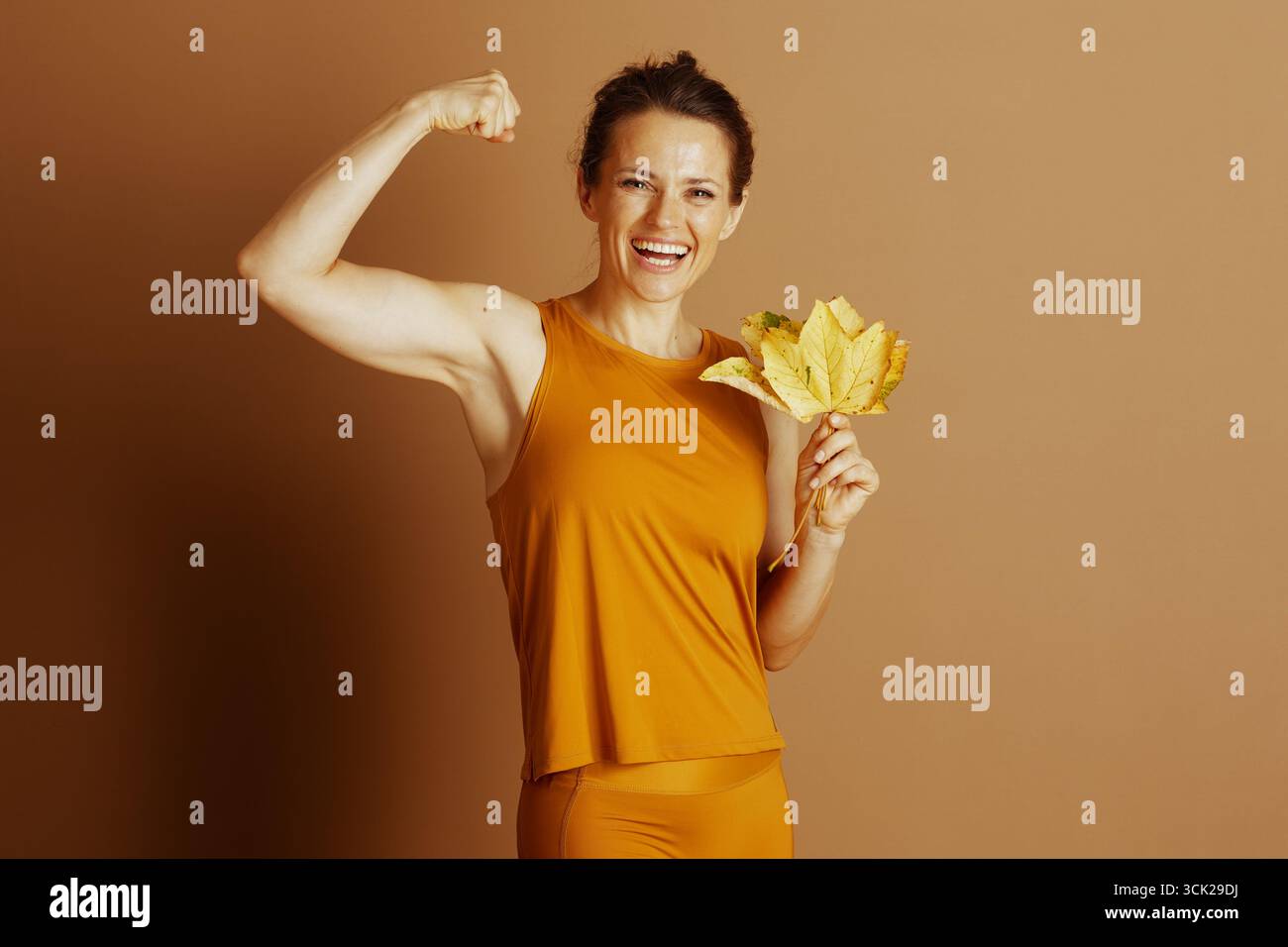 Une femme vibrante fléchit son biceps, souriant avec confiance tout en tenant des feuilles jaunes d'automne, incarnant un puissant mélange de santé, de forme physique et de saison Banque D'Images
