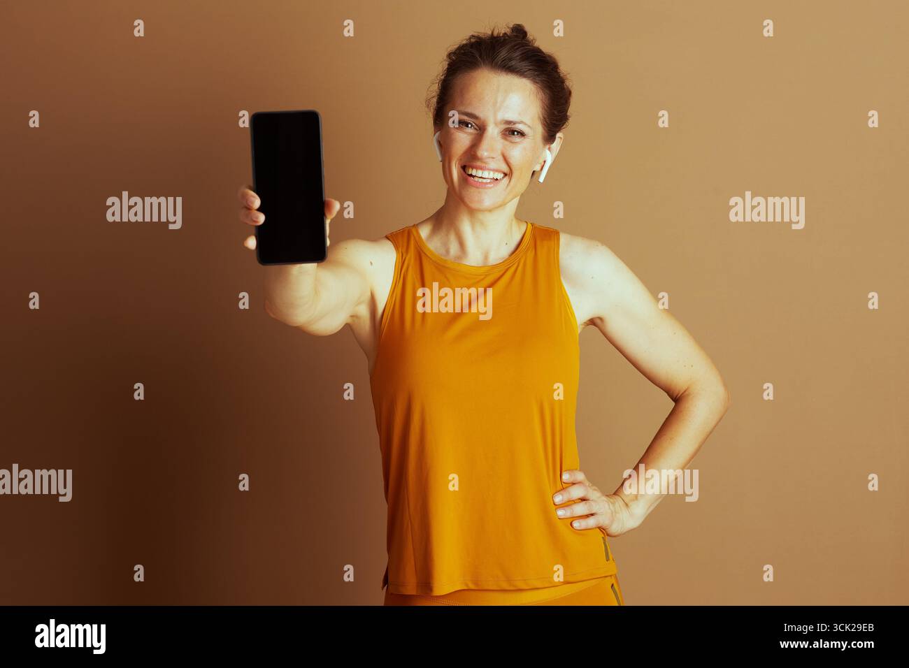 Une femme souriante en vêtements de sport jaune moutarde et des écouteurs étend un smartphone avec un écran vierge vers la caméra, main sur la hanche. Elle transmet un AC Banque D'Images
