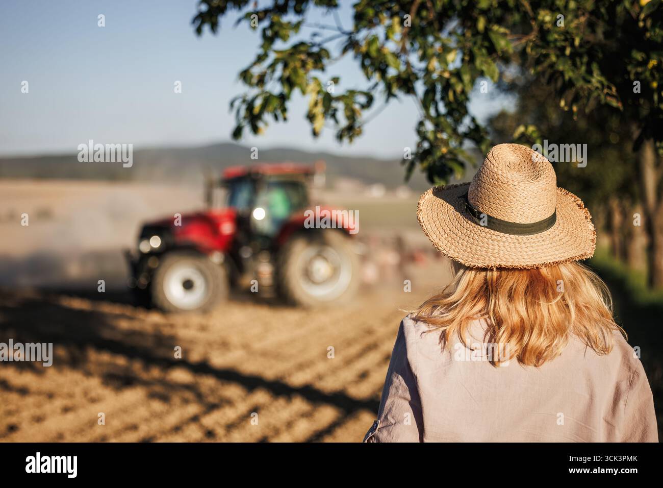 Femme fermière avec chapeau de paille regardant le tracteur avec semoir travaillant et semant la récolte sur le champ agricole Banque D'Images
