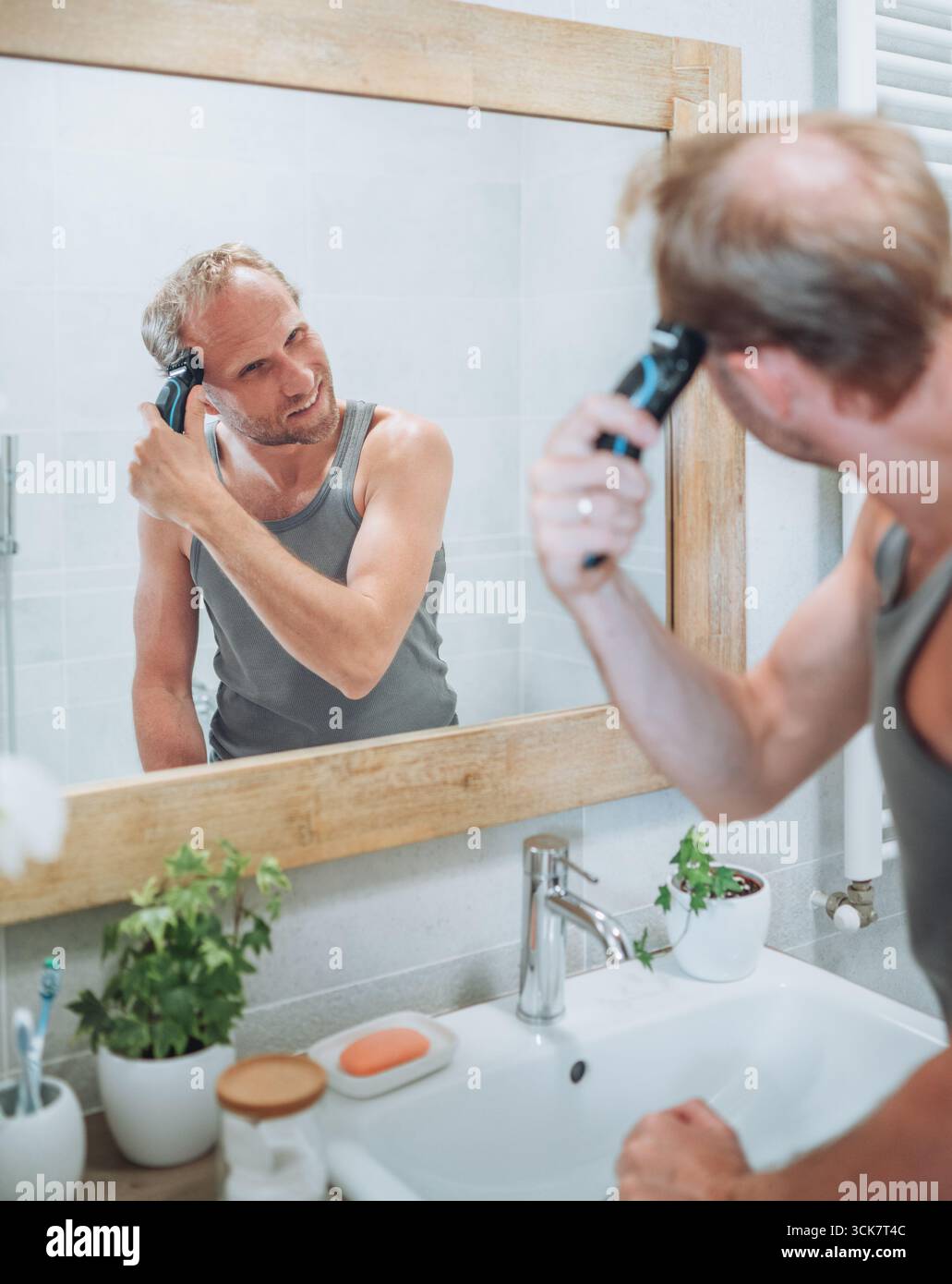 Concept corps et soin de la peau. Homme souriant se donnant une nouvelle coupe de cheveux à l'aide de tondeuse électrique rechargeable tout en regardant dans le miroir de salle de bain, Gro personnel Banque D'Images