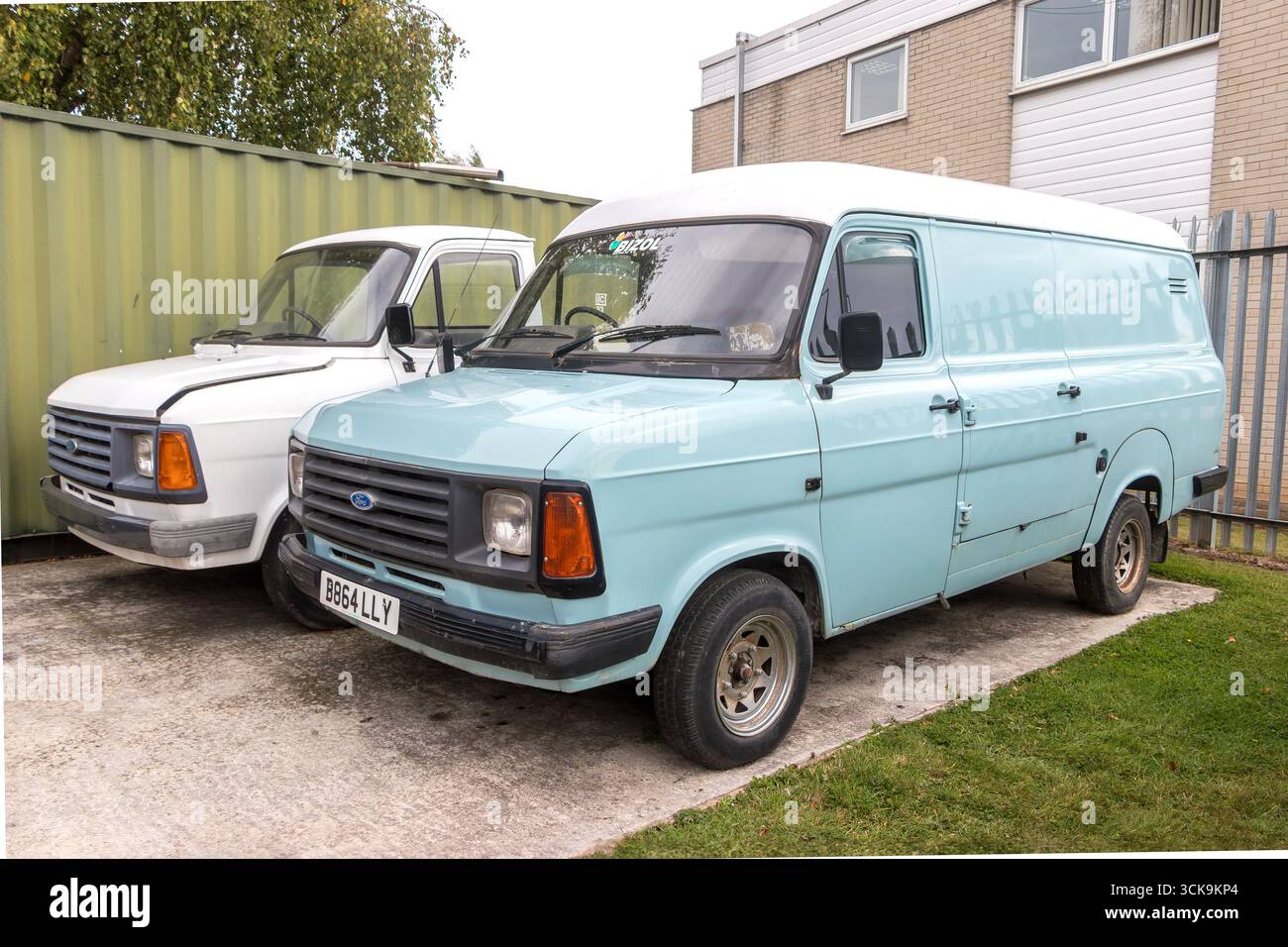 Deux fourgonnettes Ford transit vintage, une bleue et une blanche, stationnées sur une allée à côté d'un conteneur d'expédition et d'un bâtiment Banque D'Images
