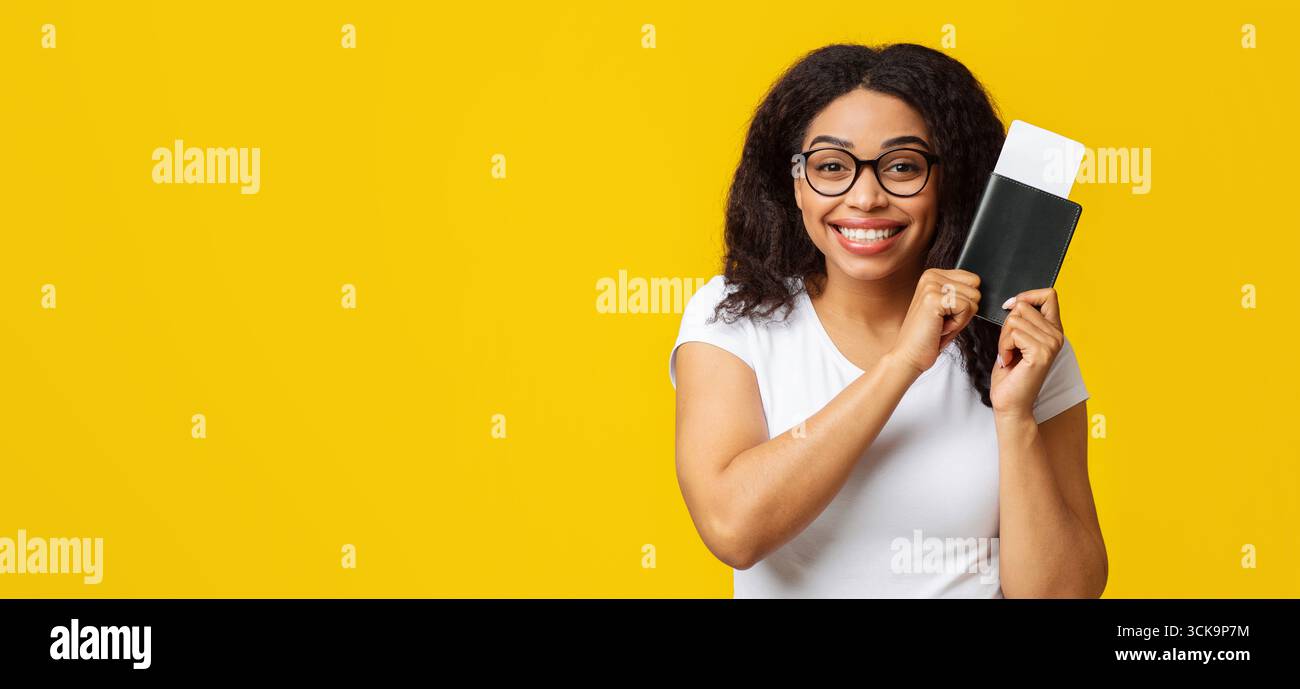 Touriste afro-américaine excitée avec passeport et billets d'avion se préparant pour les vacances, fond jaune Banque D'Images
