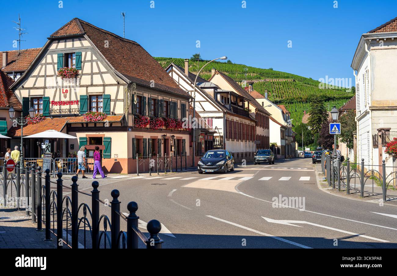 Ribeauville, France - 24 août 2025 : charmante rue du village européen avec des maisons traditionnelles à colombages, un restaurant et une colline couverte de vignobles Banque D'Images Ribeauville, France - 24 août 2025 : charmante rue du village européen avec des maisons traditionnelles à colombages, un restaurant et une colline couverte de vignobles Banque D'Images