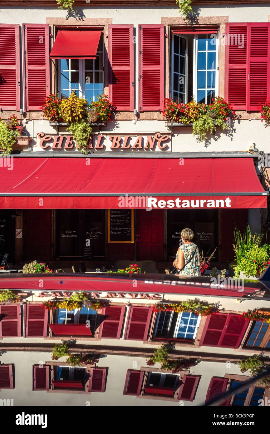 Ribeauville, France - 24 août 2025 : une charmante façade de restaurant avec des volets rouges et des fleurs, reflétant sur le toit d'une voiture, avec une femme Banque D'Images Ribeauville, France - 24 août 2025 : une charmante façade de restaurant avec des volets rouges et des fleurs, reflétant sur le toit d'une voiture, avec une femme Banque D'Images