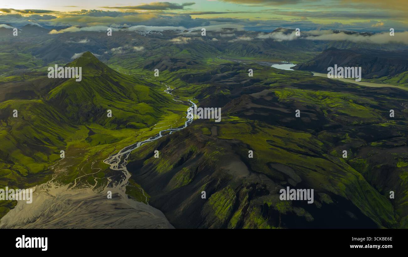 Vue aérienne d'une rivière sinueuse à travers des vallées verdoyantes et un terrain volcanique sombre, avec une montagne conique, des pentes couvertes de mousse et des sommets enneigés. Banque D'Images