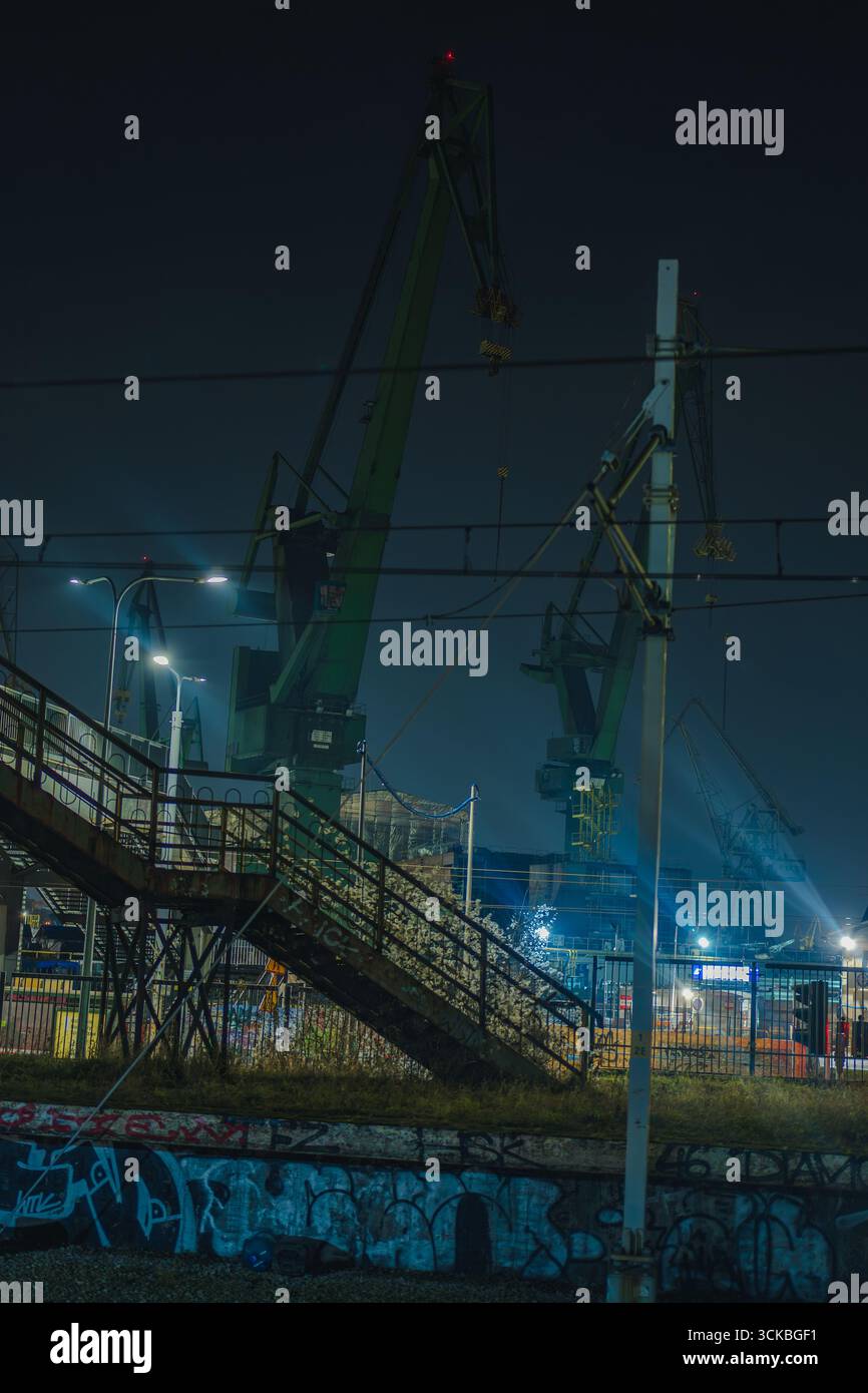 Scène nocturne Moody de grues du chantier naval de Gdansk brillant contre le ciel sombre, avec des escaliers couverts de graffitis, des voies ferrées et une faible brume au premier plan. Banque D'Images