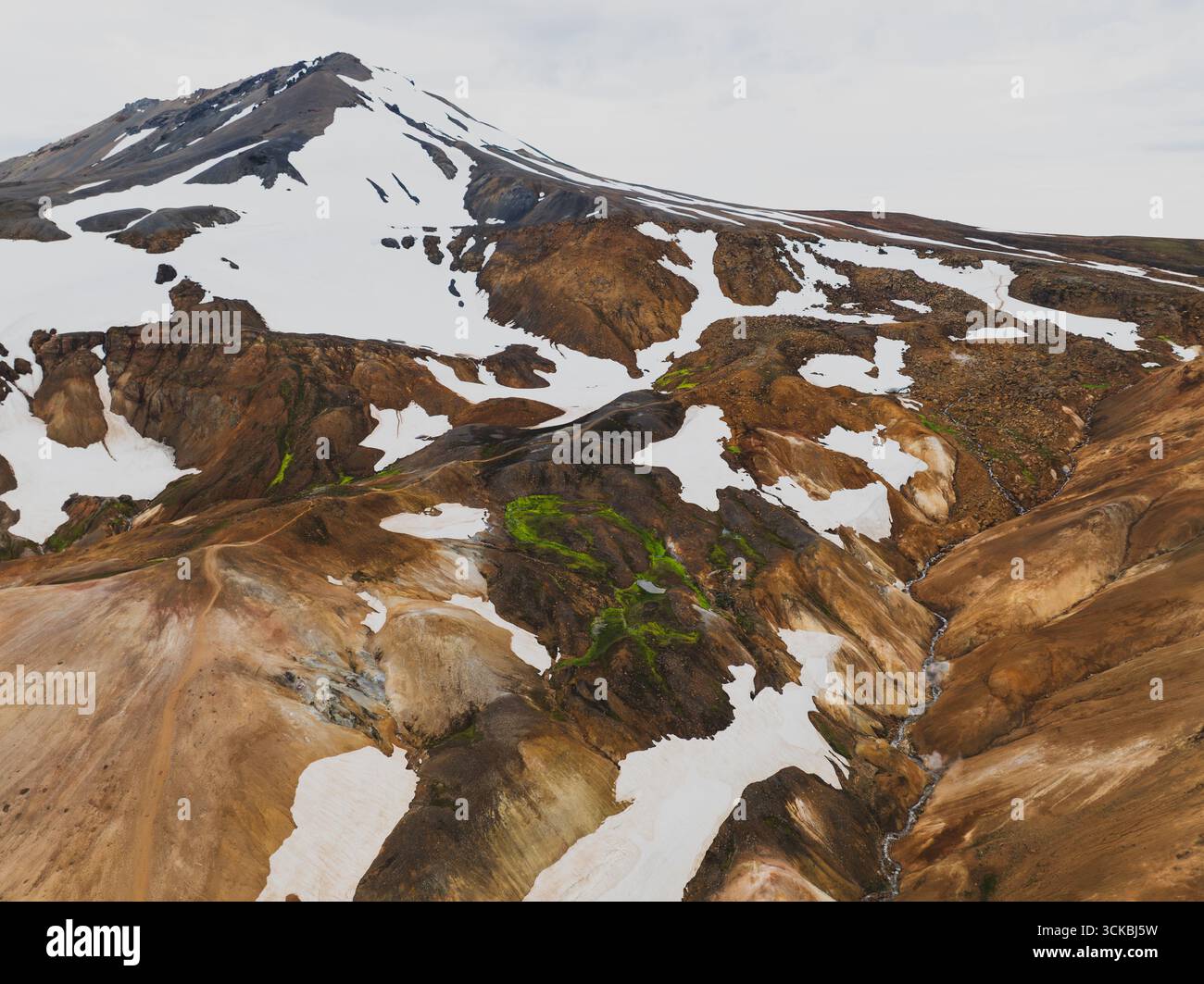 Vue aérienne d'une montagne enneigée dans les hautes terres islandaises, entourée de collines de rhyolite multicolores, de mousse verte et de tons terreux contrastés. Banque D'Images