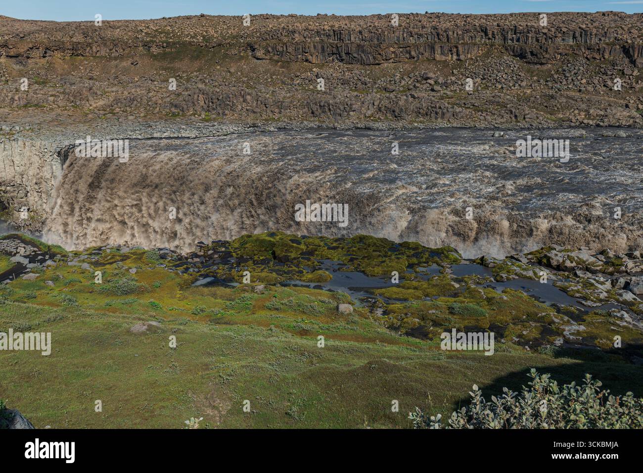 Les cascades de Dettifoss avec de l'eau boueuse au-dessus d'une falaise accidentée, entourée de roches moussues, de végétation verte et de terrains rocheux en Islande. Banque D'Images