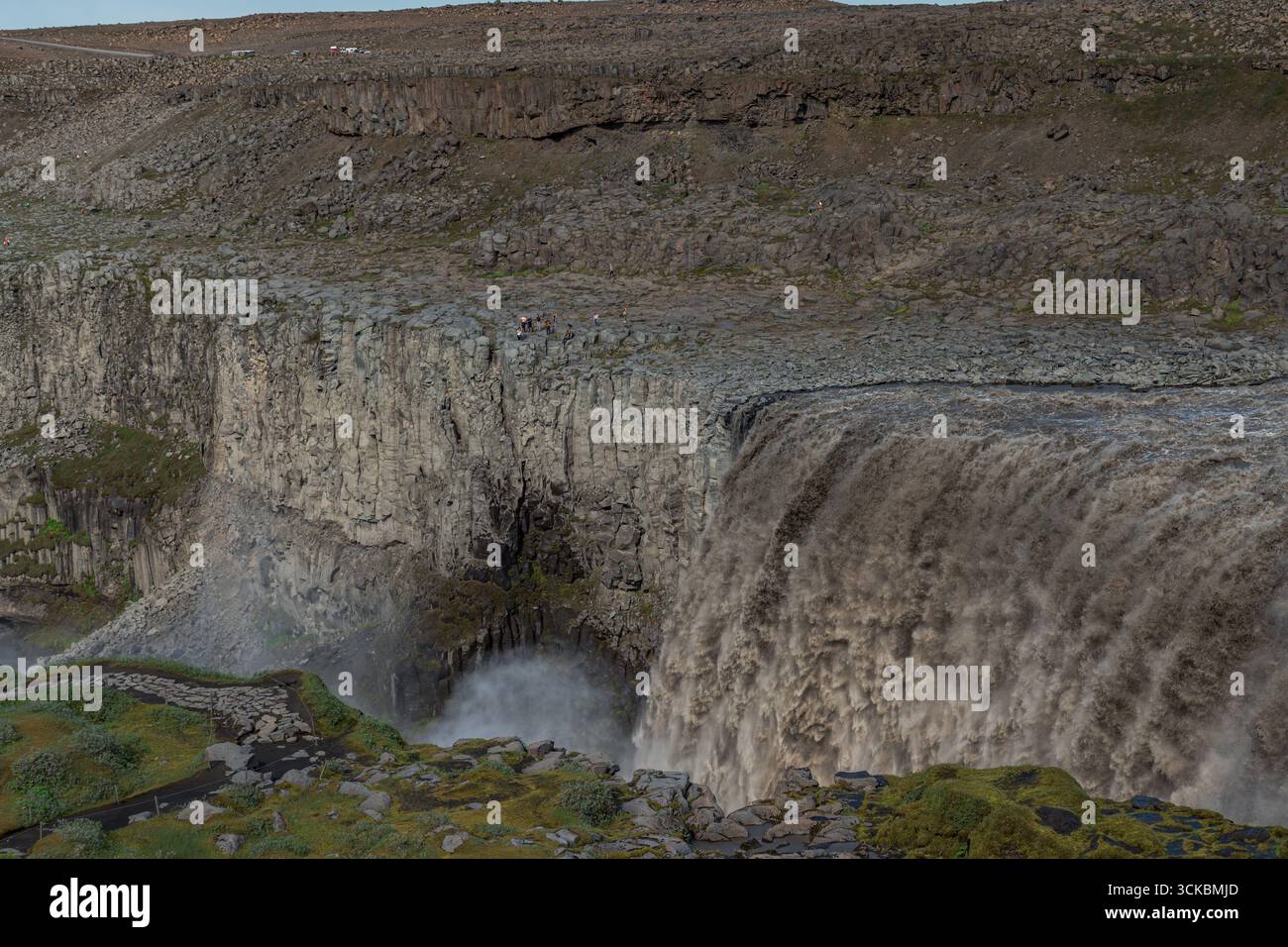 La cascade de Dettifoss cascades sur des falaises de basalte sombres, entourées d'un terrain rocheux avec des taches vertes. La brume monte comme un chemin et la zone de visualisation sont visibles Banque D'Images