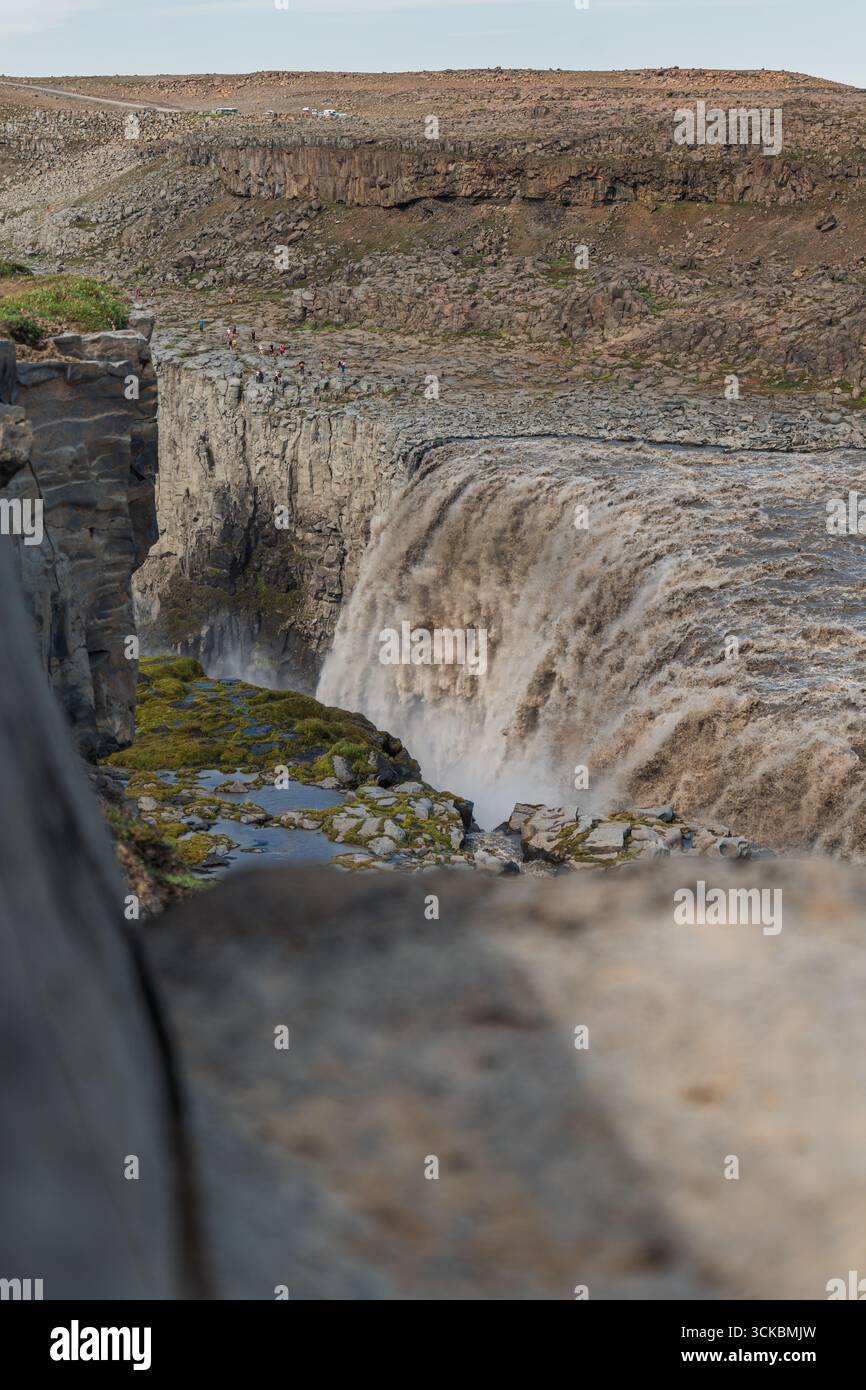Dettifoss cascades au-dessus d'une falaise accidentée avec la brume qui monte. Le terrain rocheux, la végétation clairsemée et les personnages lointains soulignent l'immense échelle de la cascade Banque D'Images