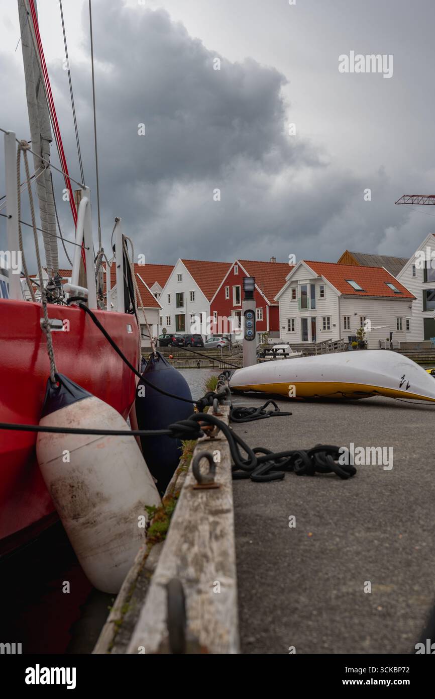 Un voilier rouge avec des ailes visibles et des lignes d'amarrage repose sur un quai. A proximité se trouvent des maisons aux toits blancs et rouges, avec des nuages gris au-dessus dans une Norvégie Banque D'Images