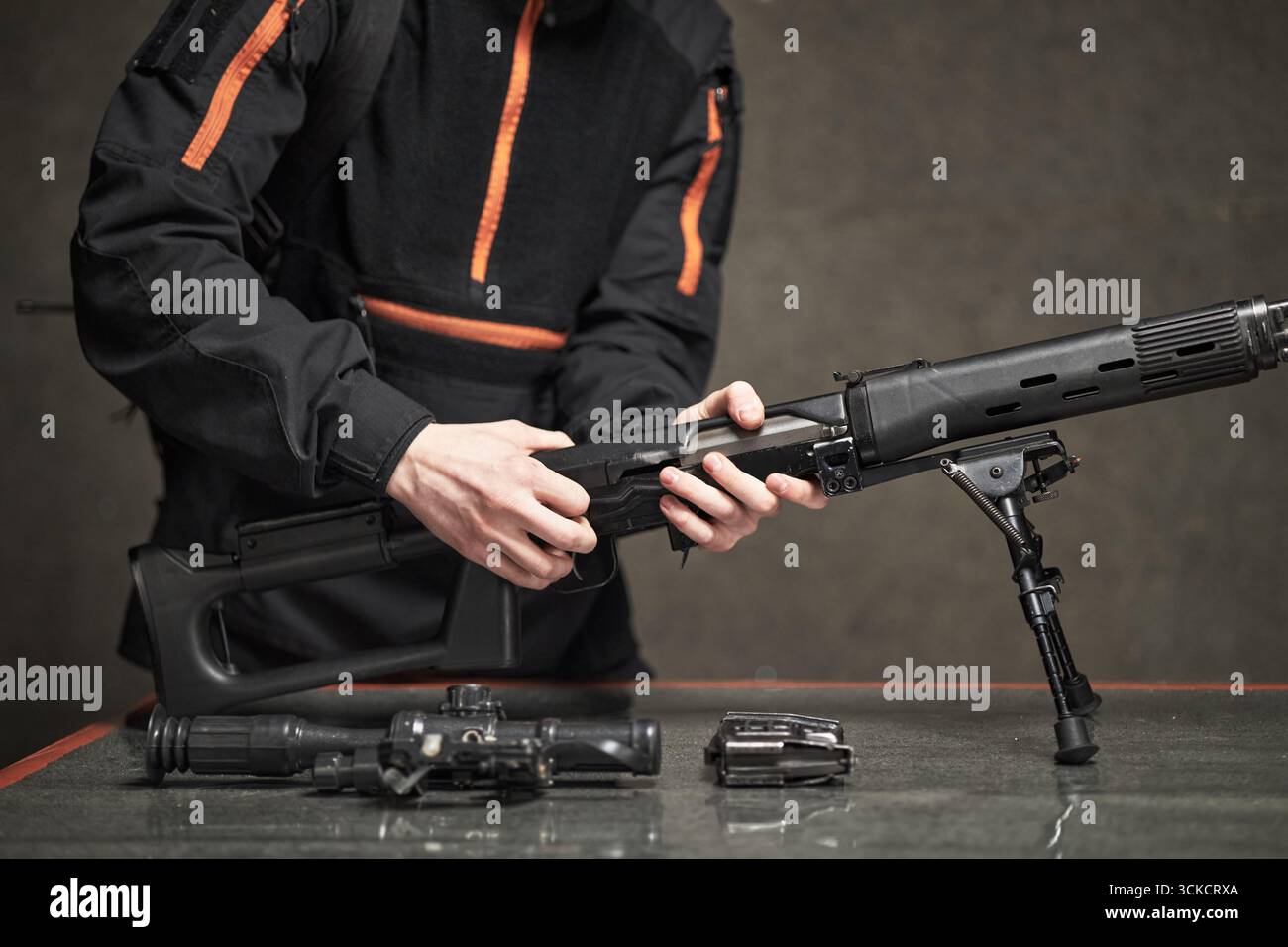 Jeune homme caucasien adulte assemblant un fusil d'assaut sur la table, portant une veste tactique, les mains ajustant les composants de l'arme à feu, des accessoires d'arme supplémentaires placés à proximité Banque D'Images